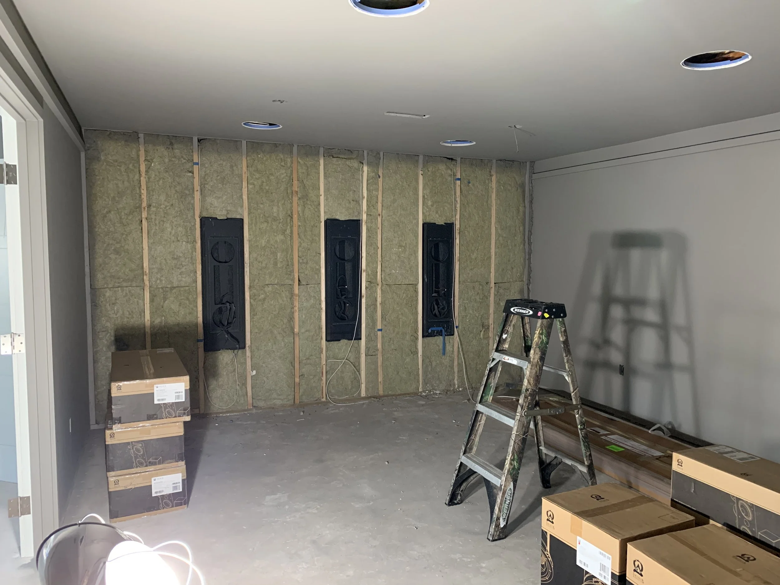Atmos theater room under renovation with exposed wall insulation, black electrical panels, a stepladder, and boxes of supplies on the floor.