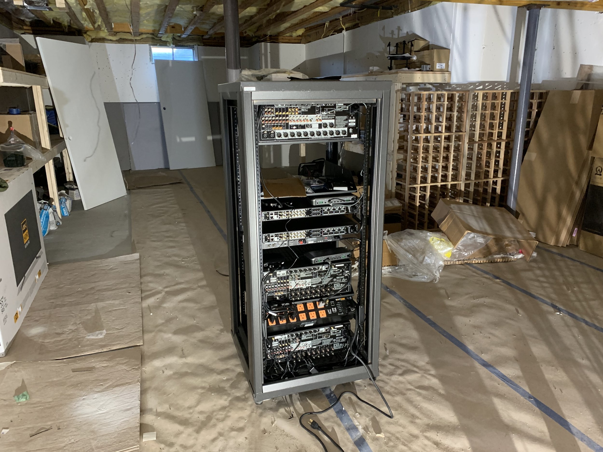 An equipment rack filled with electronic audio or video gear is in the center of a basement or storage room with exposed insulation and unfinished walls. Surrounding the rack are shelves, cardboard, and packaging materials.