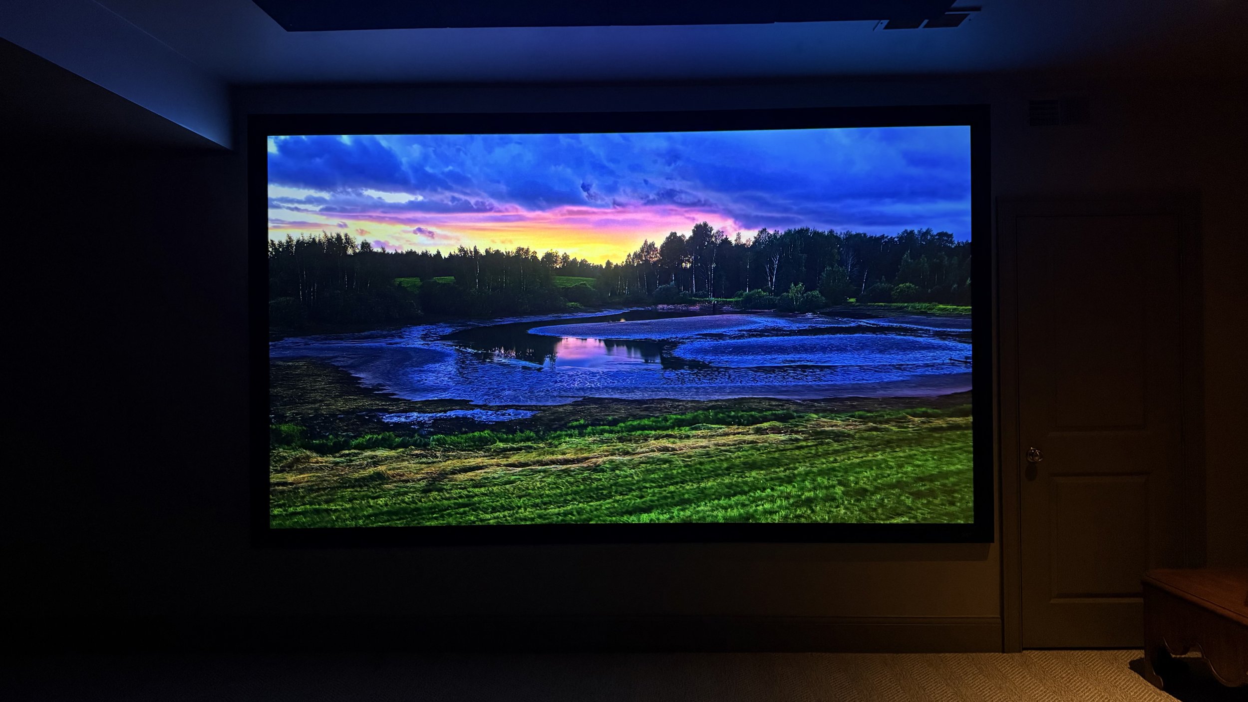 Laser projector and large screen displaying a landscape of a river with trees and a colorful sunset sky.