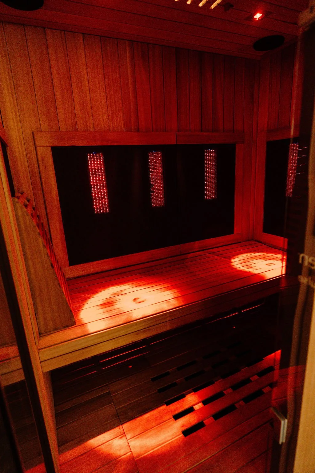 Inside a wooden sauna with bench seating, red LED lights reflecting on the wood, and a light pattern on the floor.