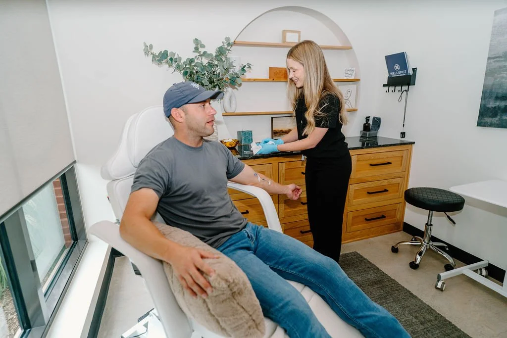 A man at a medical clinic receiving an IV infusion while a nurse prepares to administer the treatment. The man is sitting in a reclining chair near a window, and the nurse is standing beside him, smiling. The room has a wooden cabinet, shelves with decor, and medical equipment.