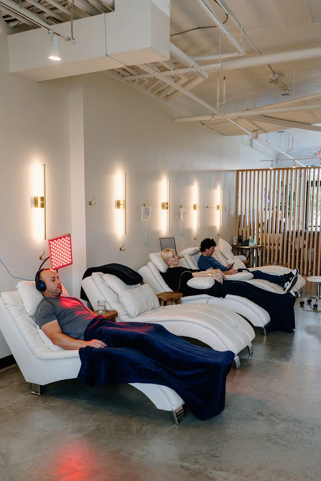 People resting on recliner chairs in a modern lounge with white walls, wooden accents, and soft lighting, in a hospital or wellness center setting.