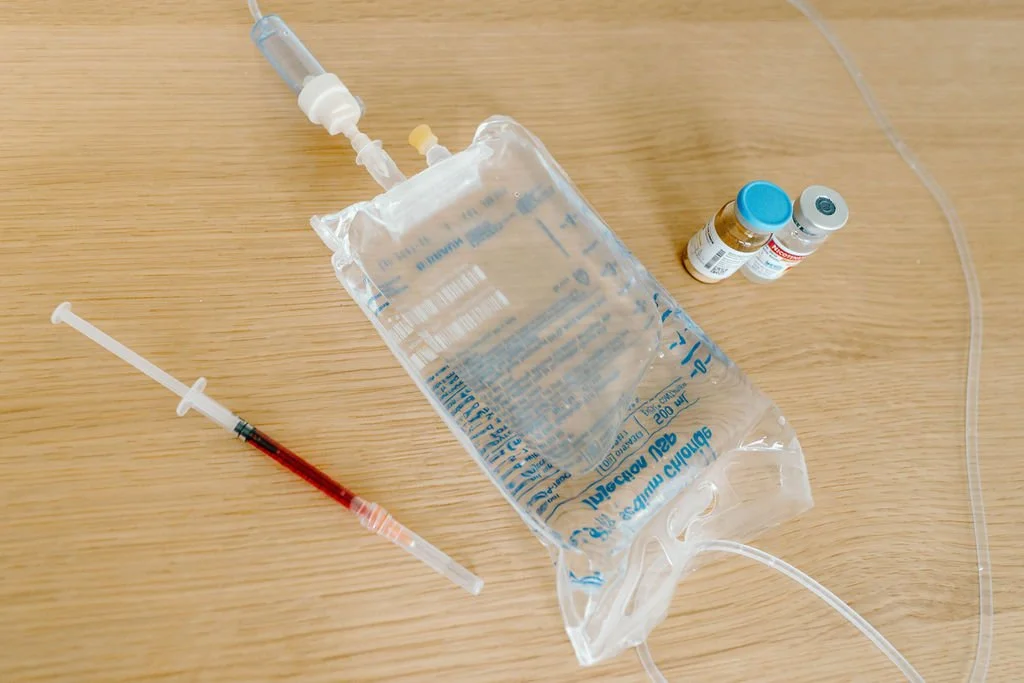 Medical supplies on a wooden surface, including a IV fluid bag, a syringe with blood, and two small vials.