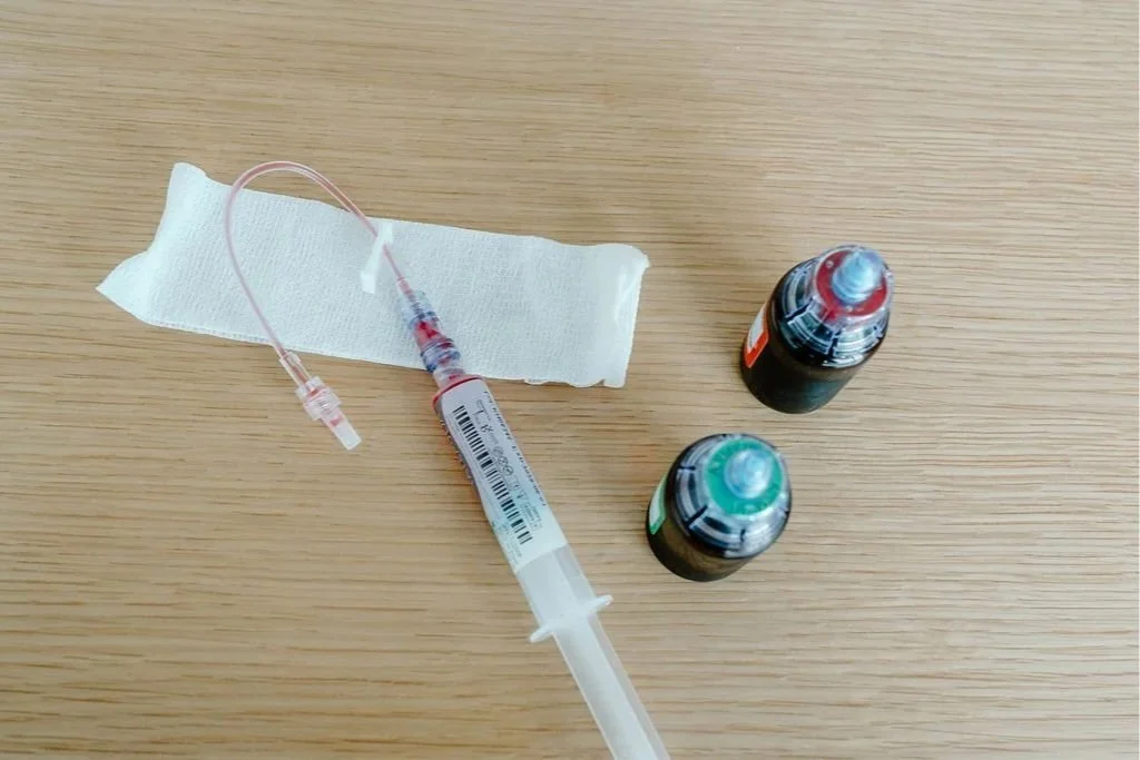 A syringe with tubing, a piece of gauze, and two vials of medication on a wooden surface.