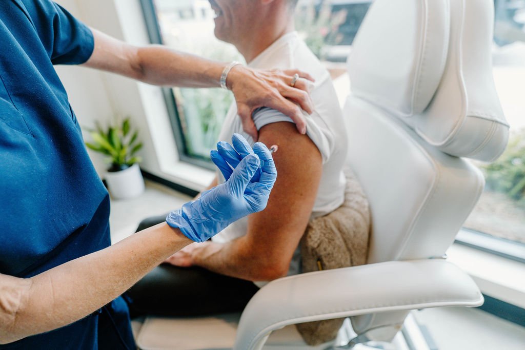A person receiving a vaccination shot in their upper arm while seated in a white chair, with a healthcare professional wearing blue gloves preparing to give the shot, in a bright room with large windows and a potted plant in the background.