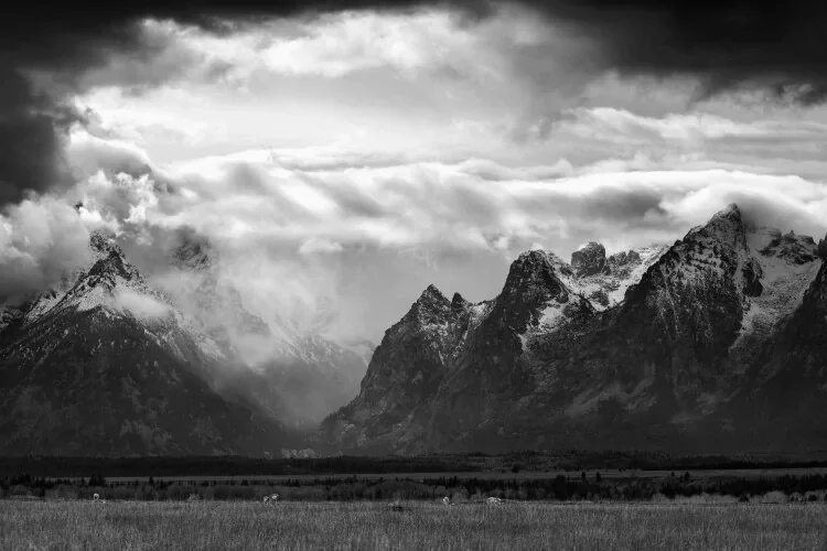 Black and white photograph of rugged mountain range under cloudy sky, with a flat foreground.