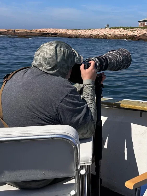 A person sitting on a boat with a camouflage hoodie, holding a large camera with a long telephoto lens aimed at the water.