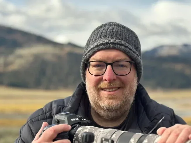 Man smiling outdoors wearing glasses, a gray beanie, and a black jacket, holding a camera with a telephoto lens.