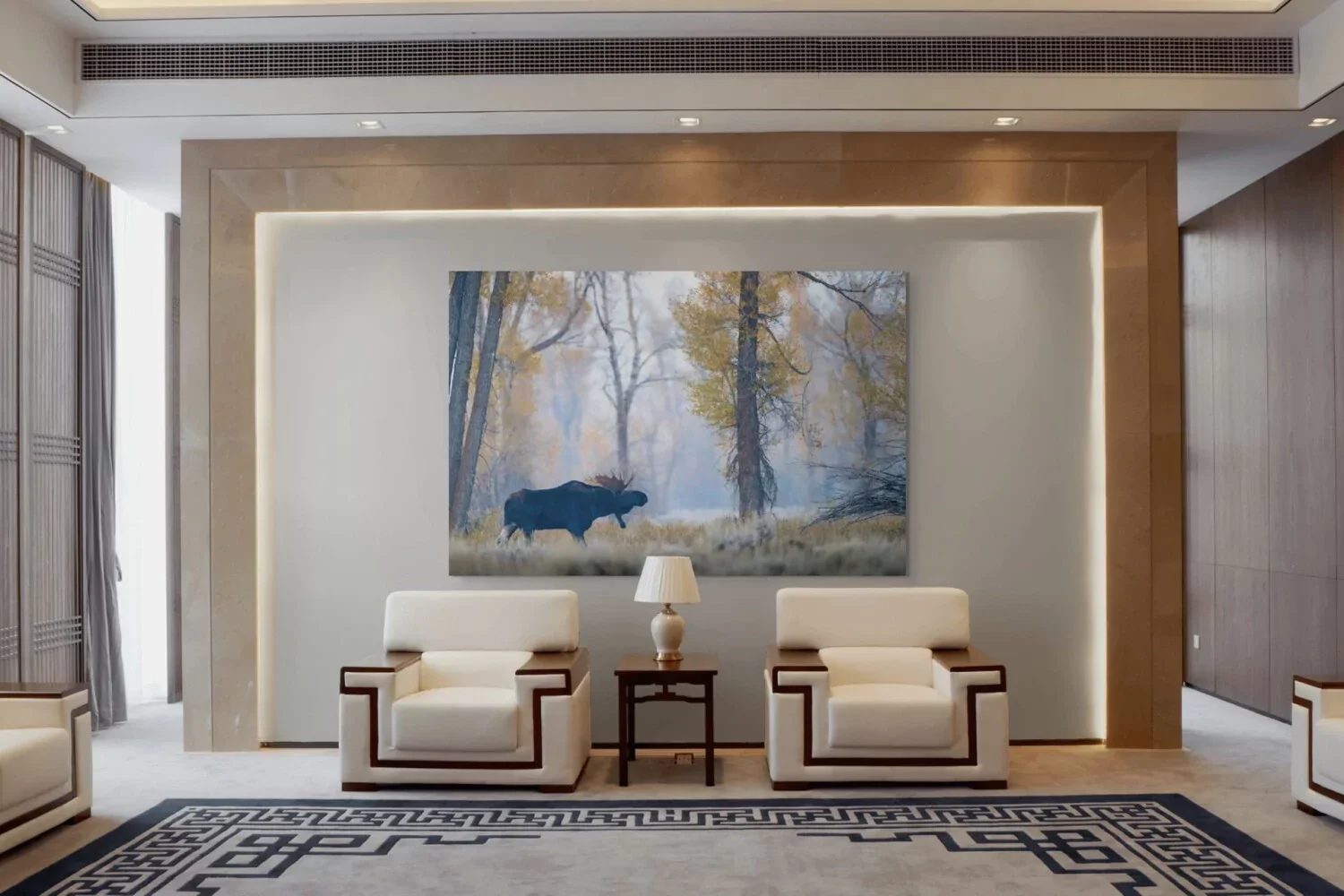 Modern hotel lobby with two white armchairs, a small wooden table with a lamp, a large painting of a bear in a forest, and a patterned rug.