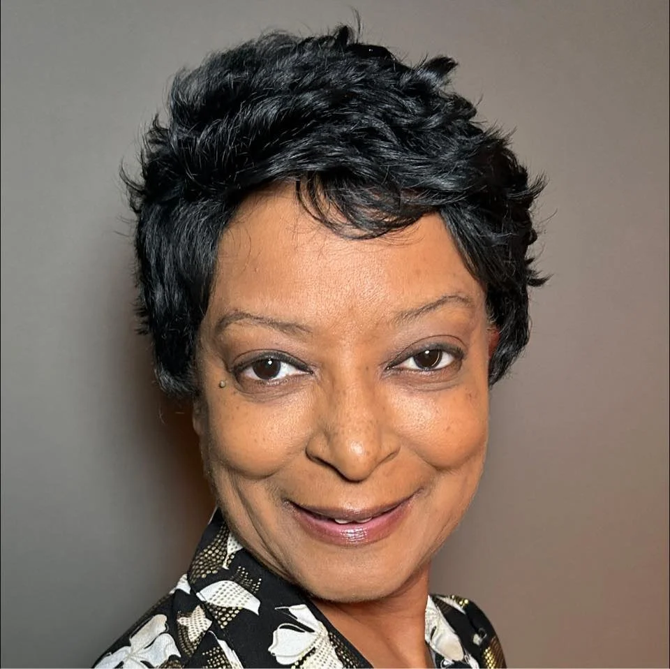 A woman with short black hair, smiling, wearing a black and white patterned top, standing against a plain background.