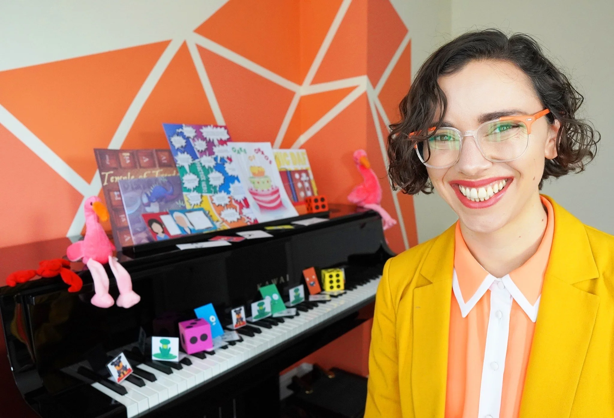 A smiling woman with short curly hair and glasses wearing a yellow blazer, standing next to a black piano decorated with colorful paper dice and plush toys, in a room with orange geometric wall art.