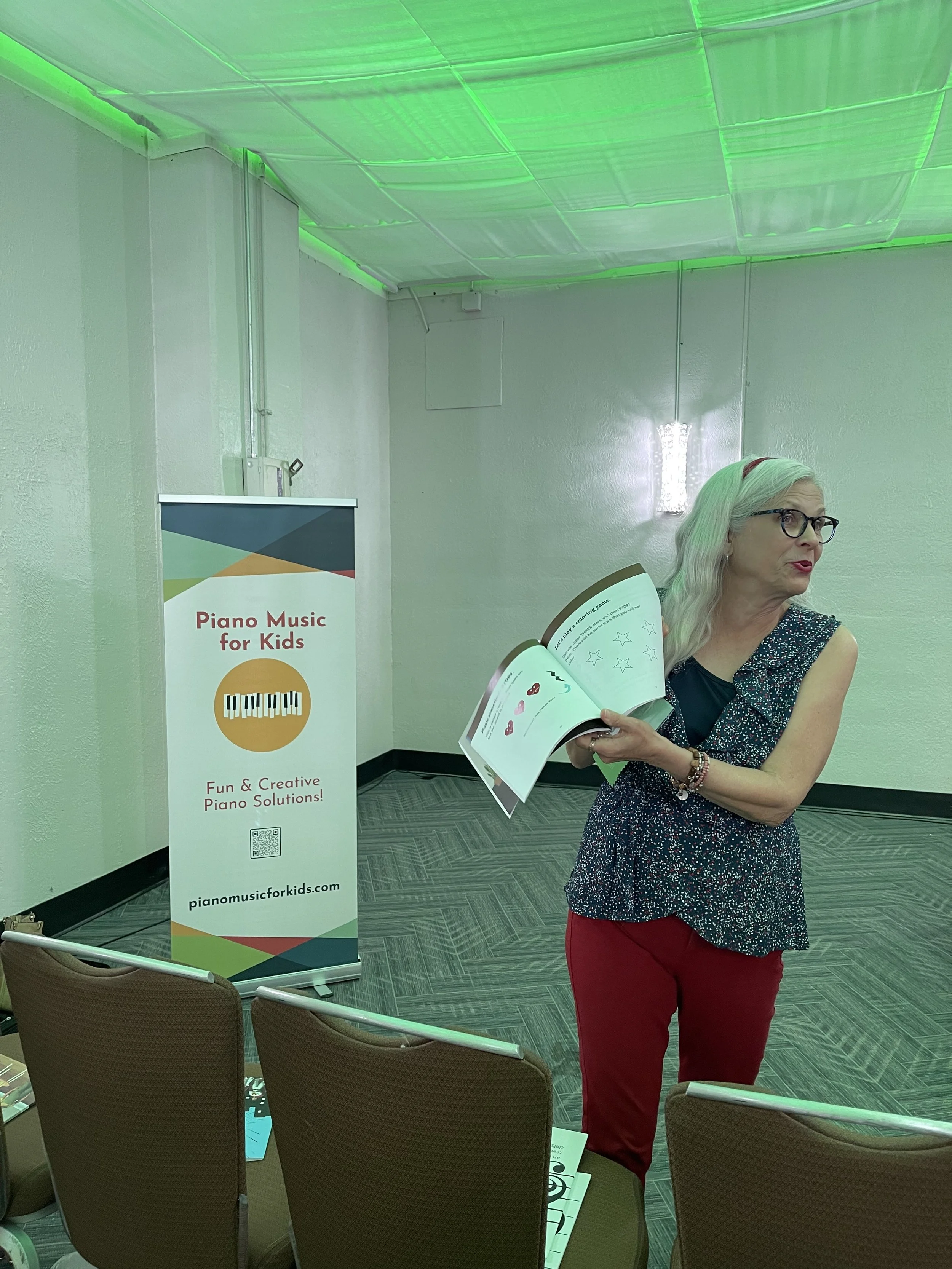 Woman with gray hair, glasses, and a red headband holding a sheet of paper at a piano music for kids presentation. Sign next to her reads 'Piano Music for Kids' and 'Fun & Creative Piano Solutions!'