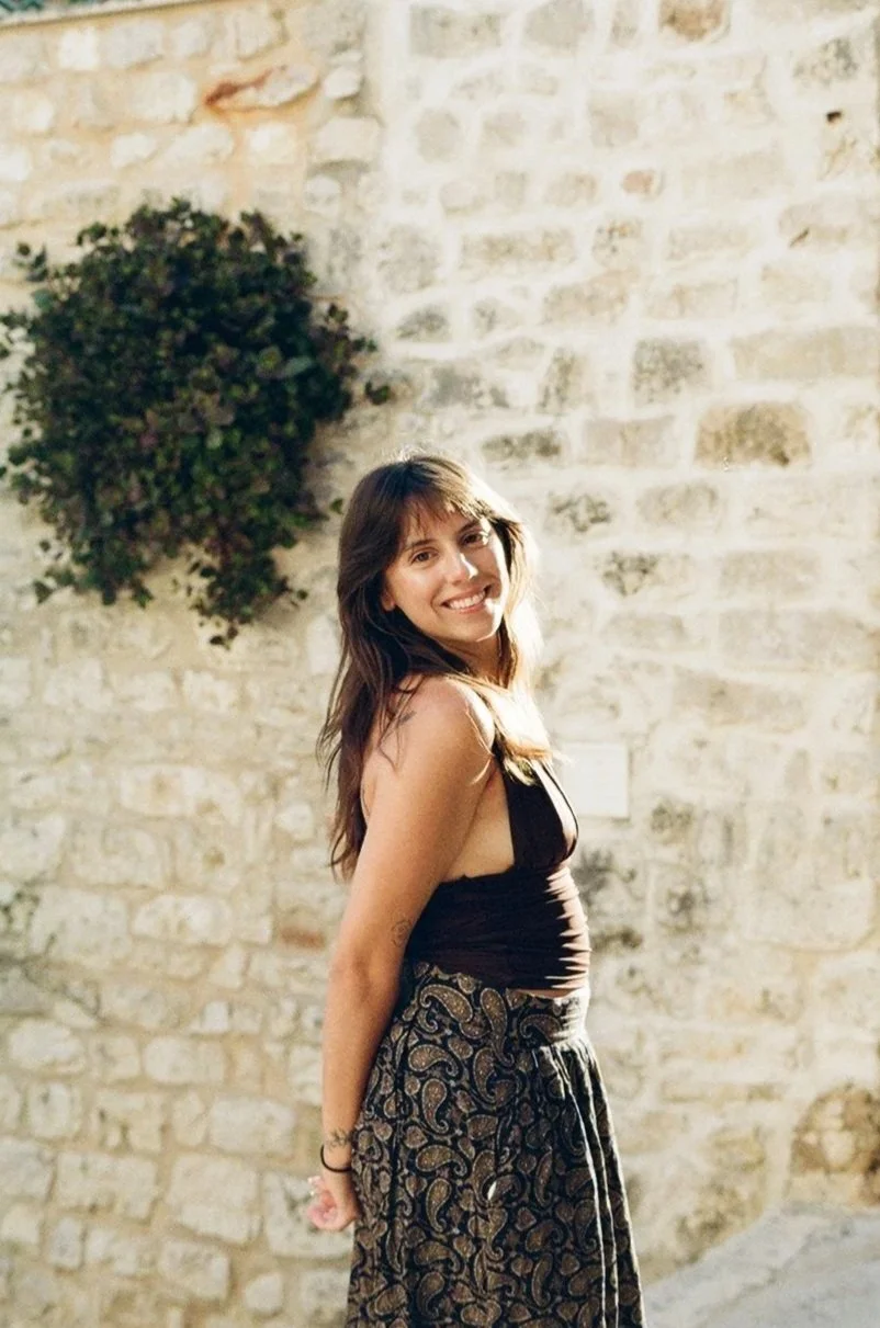 A woman with long brown hair smiling and facing slightly to the right, standing outdoors in front of a stone wall and a hanging plant.