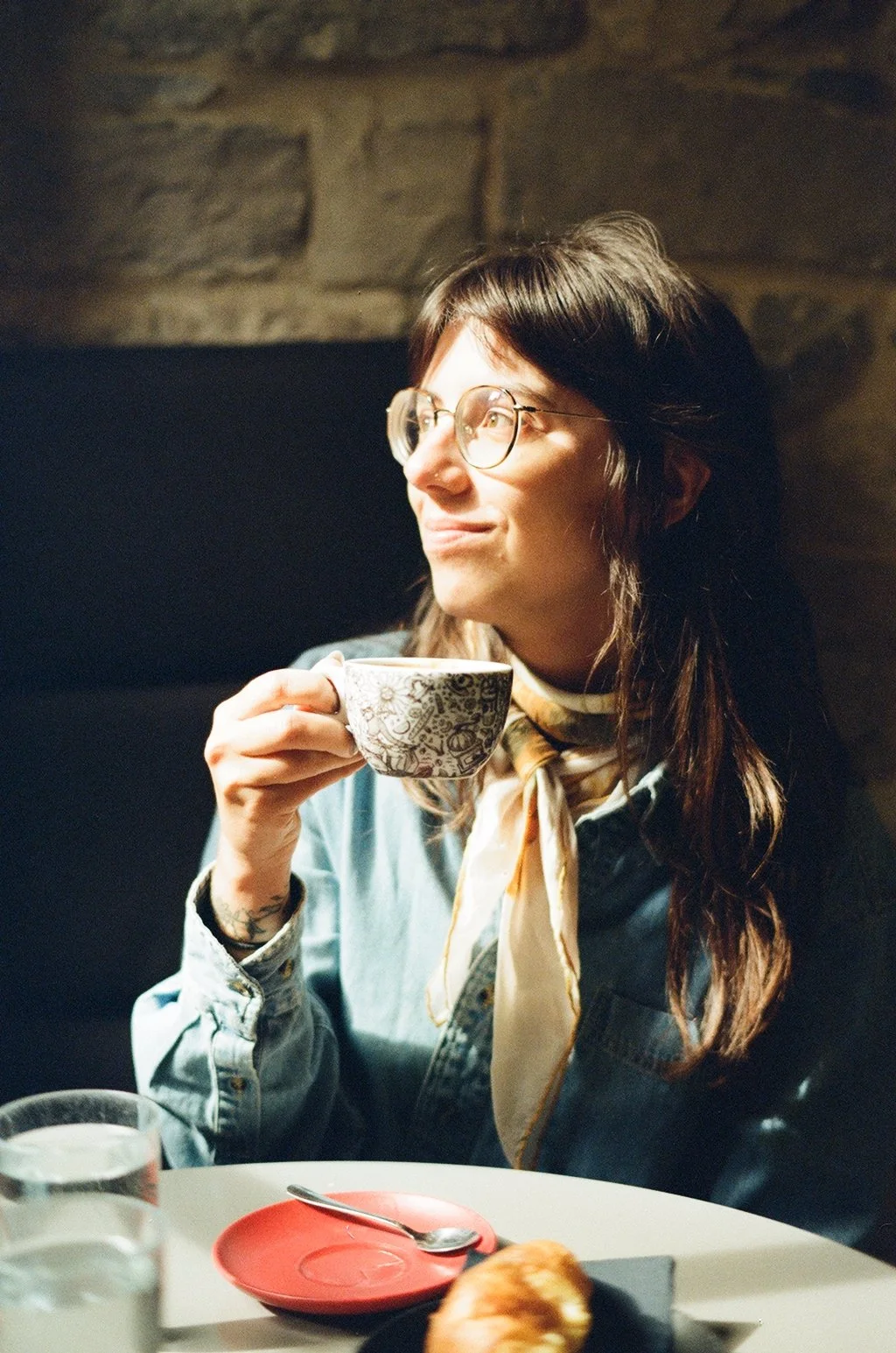 A young woman with glasses and long brown hair, wearing a denim shirt and scarf, sitting at a table with a cup of coffee or tea, smiling and looking to her left.