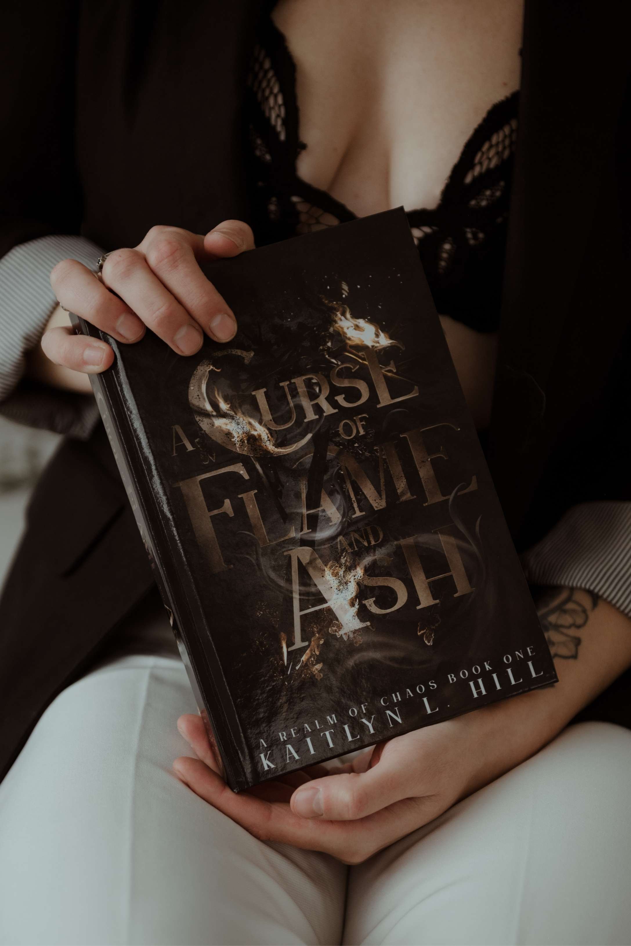 Close-up of hands holding the novel A Curse of Flame and Ash by Kaitlyn L. Hill, with the torso softly blurred in the background.
