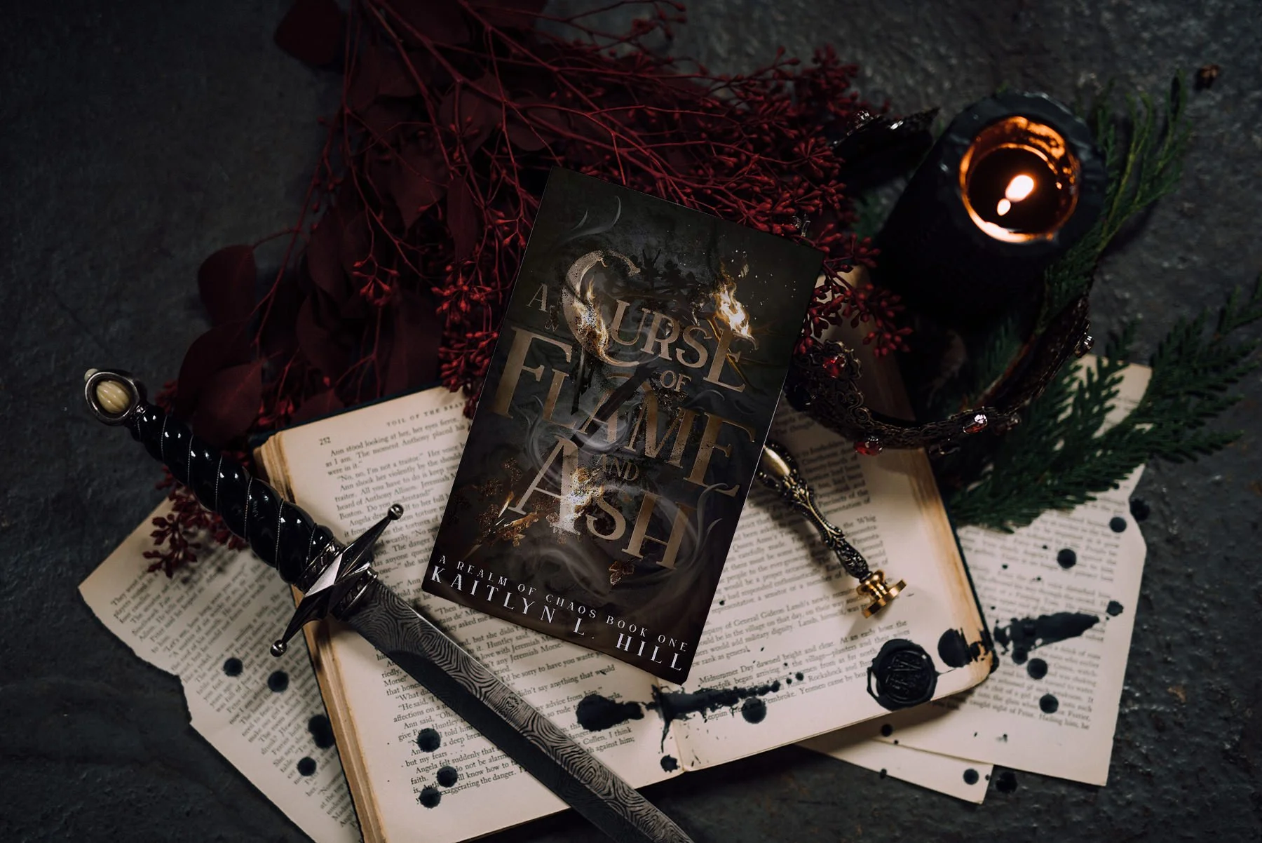 Flatlay featuring the novel A Curse of Flame and Ash, Book One in the A Realm of Chaos series by author Kaitlyn L. Hill, surrounded by a dagger, spilled ink, a metal crown, a candle, and deep red florals in a dark fantasy aesthetic.