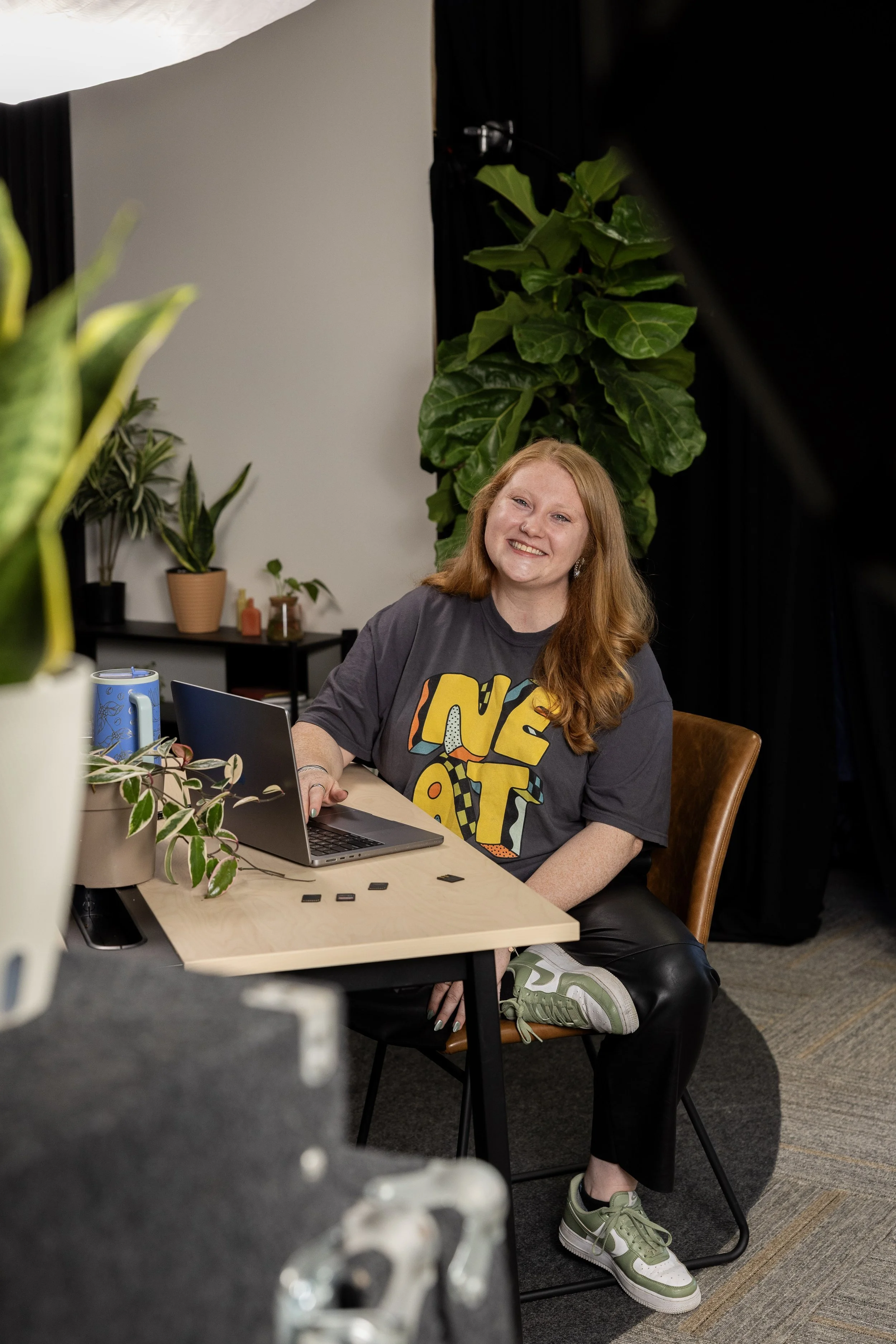 Smiling red-haired woman working on a laptop at a plant-filled desk.