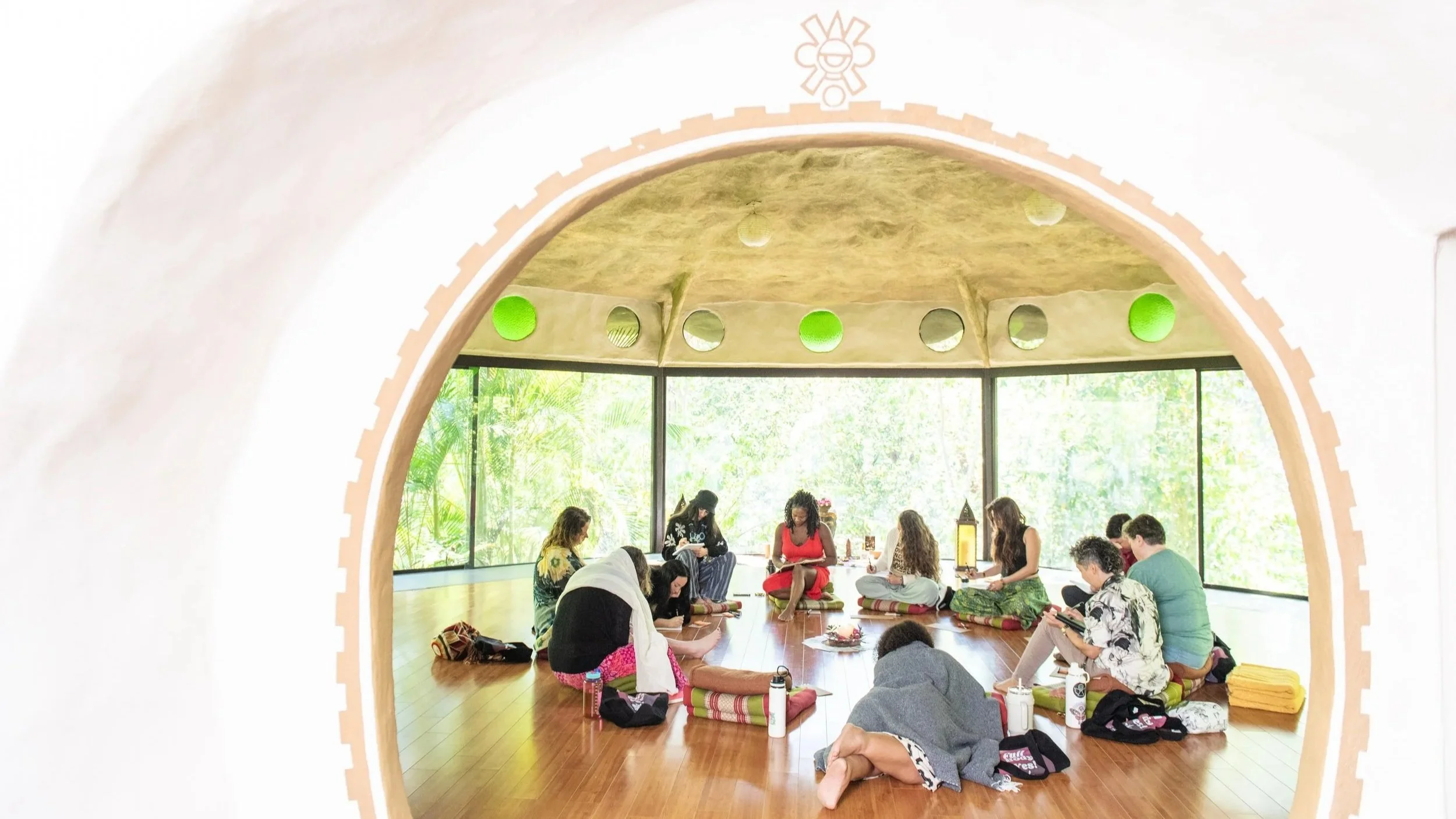 A group of people sitting on the floor in a semi-circle during a meditation or yoga session inside a spacious room with large windows and green foliage outside. retreat photographer Briana Cerezo
