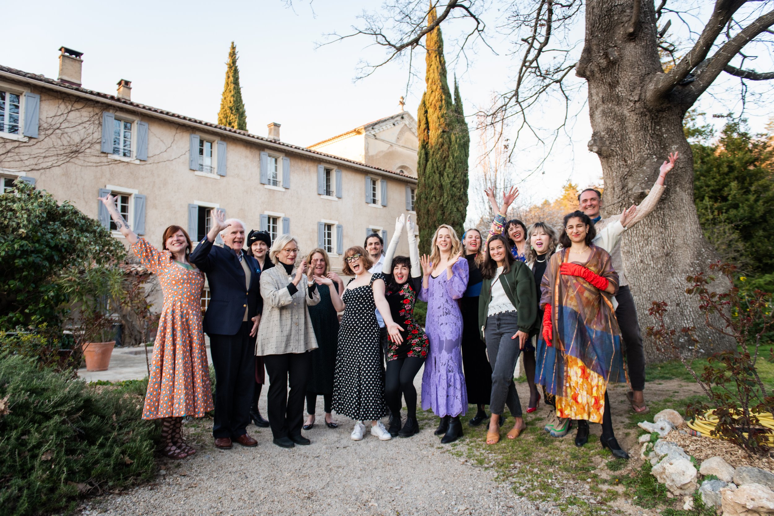 A group of diverse people smiling and raising their hands outdoors in front of a large tree and a beige building with blue shutters. trauma-informed breathwork guide Portland Oregon