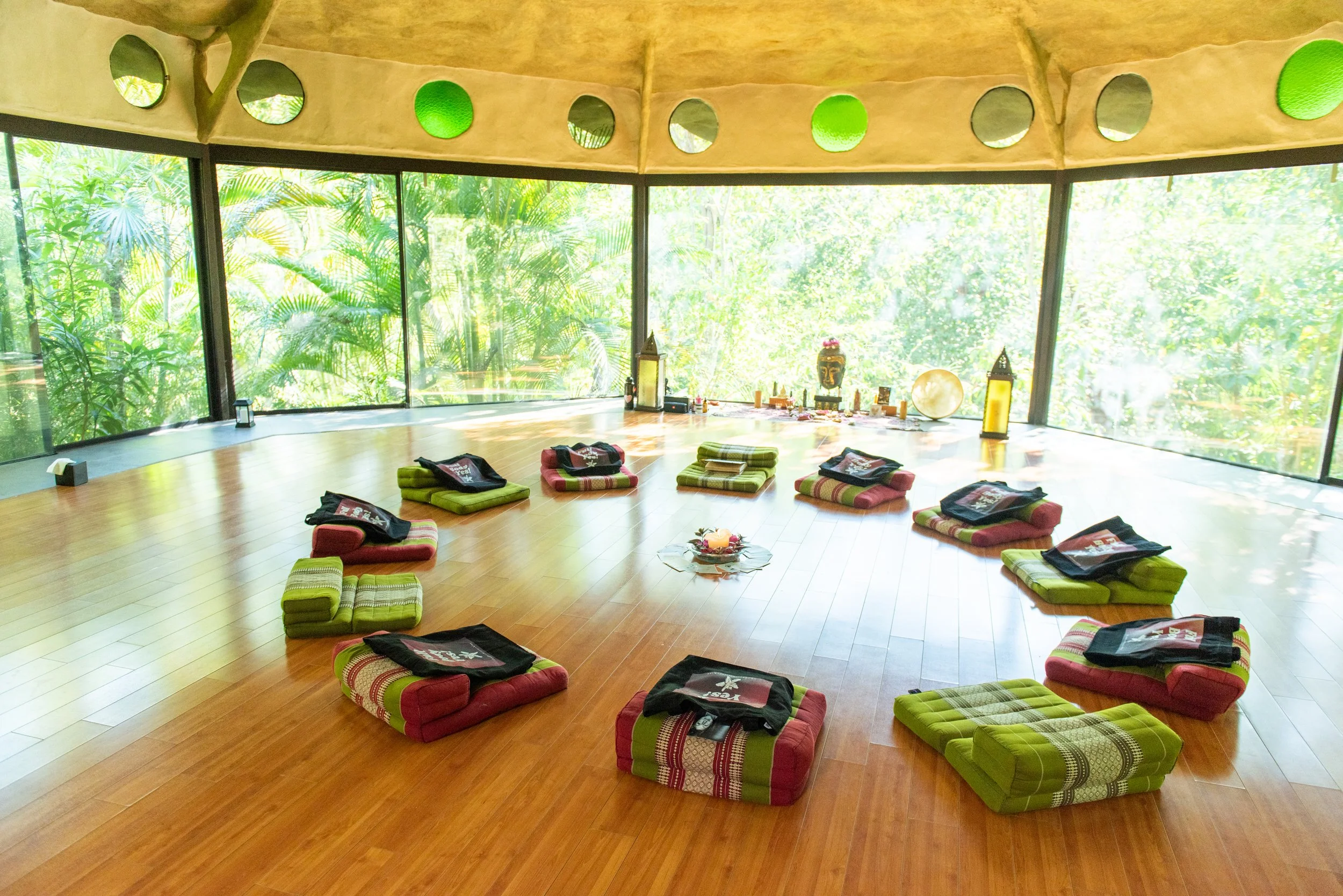 A serene meditation room with large glass windows revealing lush green tropical foliage outside. The wooden floor is arranged with folded cushions and bolsters for seating, along with small decorative items, lanterns, and a Buddha face statue, creati