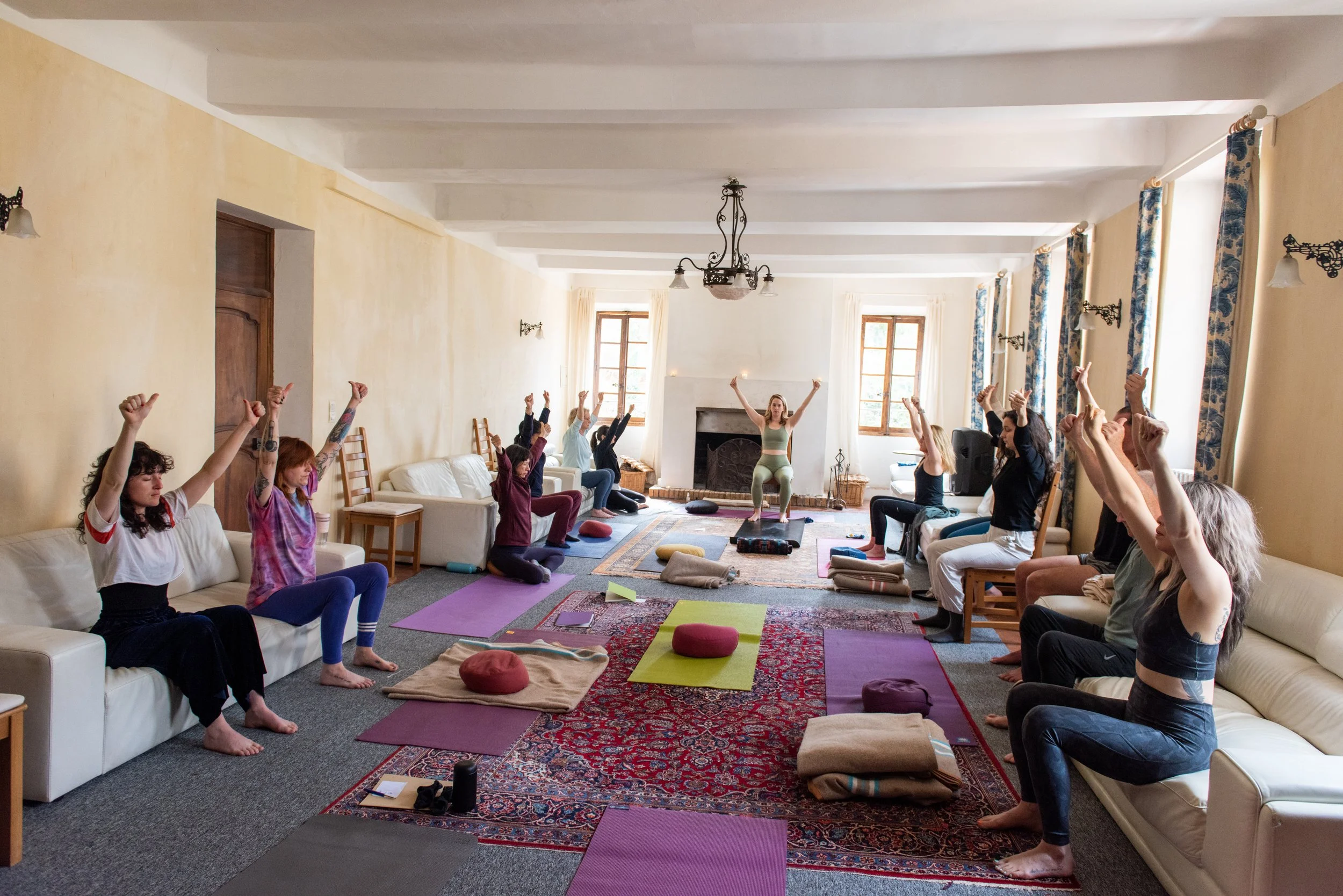 People participating in a yoga or meditation session in a bright, spacious room with large windows, colorful mats, cushions, and a fireplace. retreat facilitator