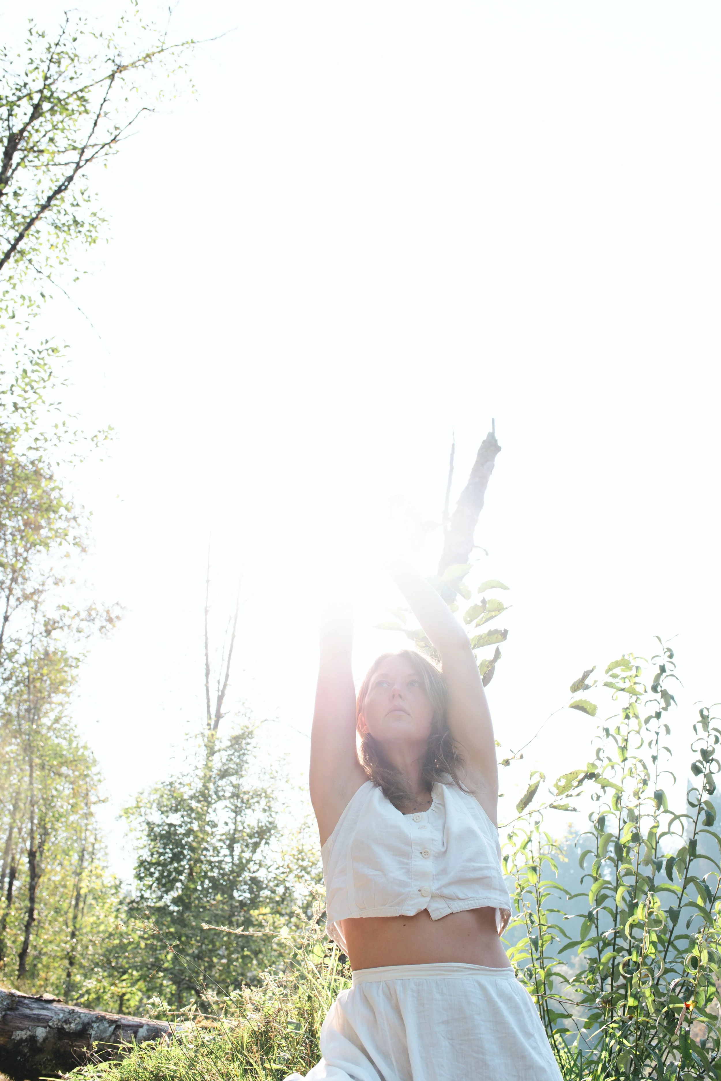 Briana Lynn Cerezo in a white sleeveless top and white pants standing outdoors in a lush green forest, raising her arms toward the bright sunlight. Breathwork coach portland