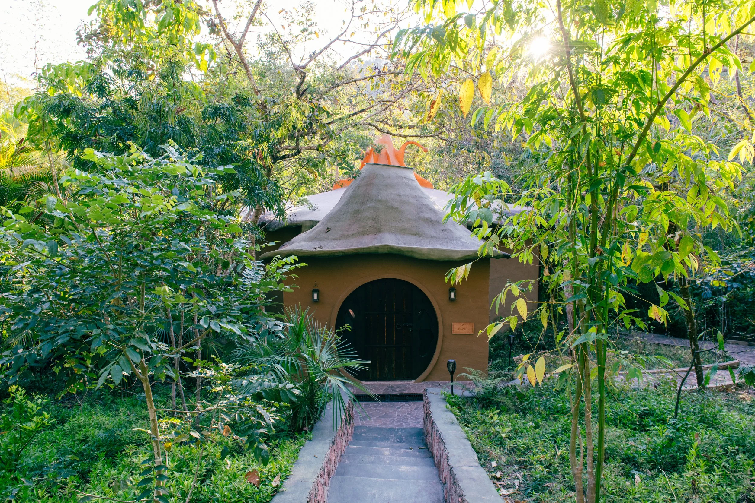 A small house or cottage with a rounded door, surrounded by lush green foliage and trees, with sunlight shining through the leaves. breathwork event portland