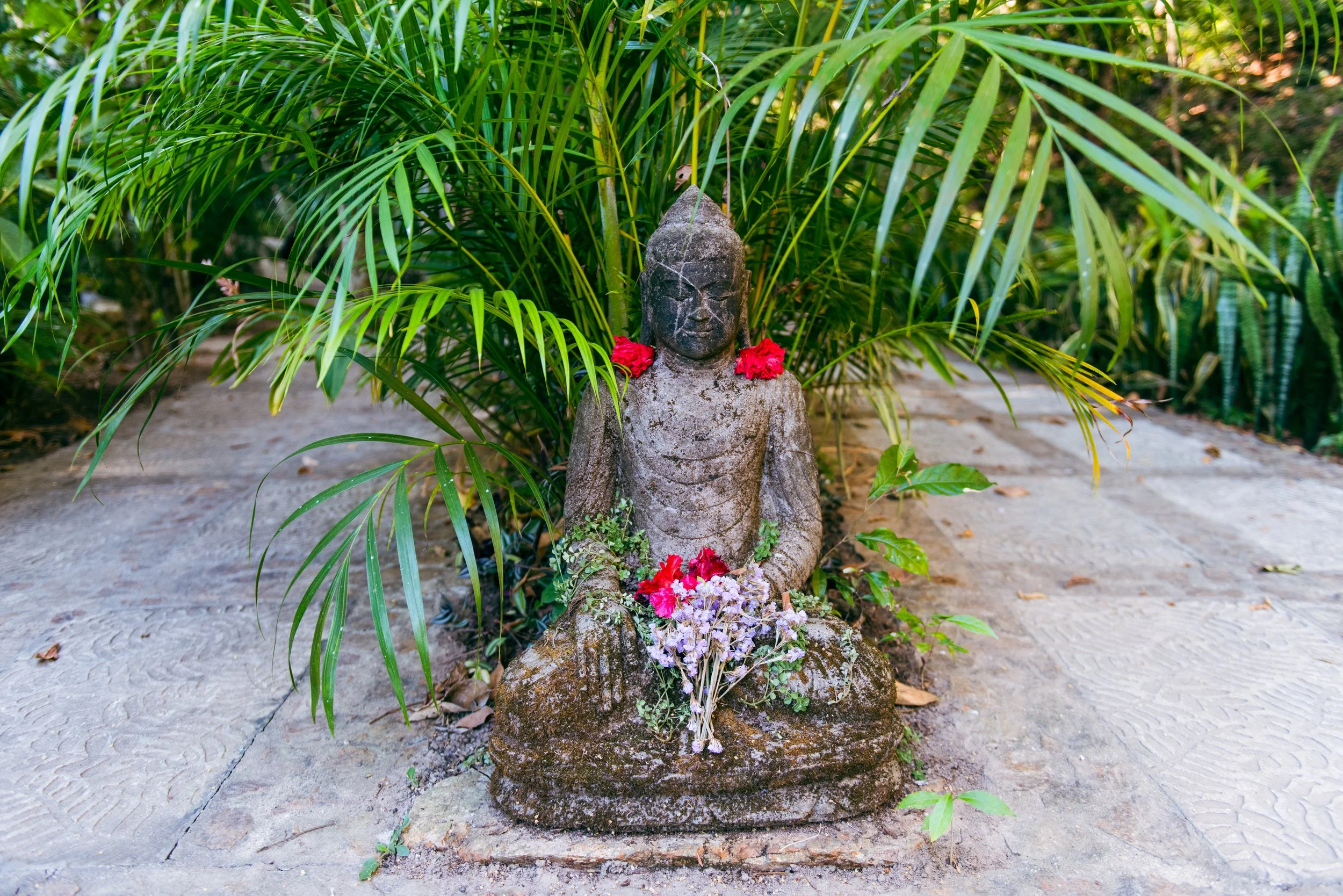 Stone statue with a calm face, surrounded by red and purple flowers, situated on a sandy ground with lush green foliage in the background. Breathwork guide portland