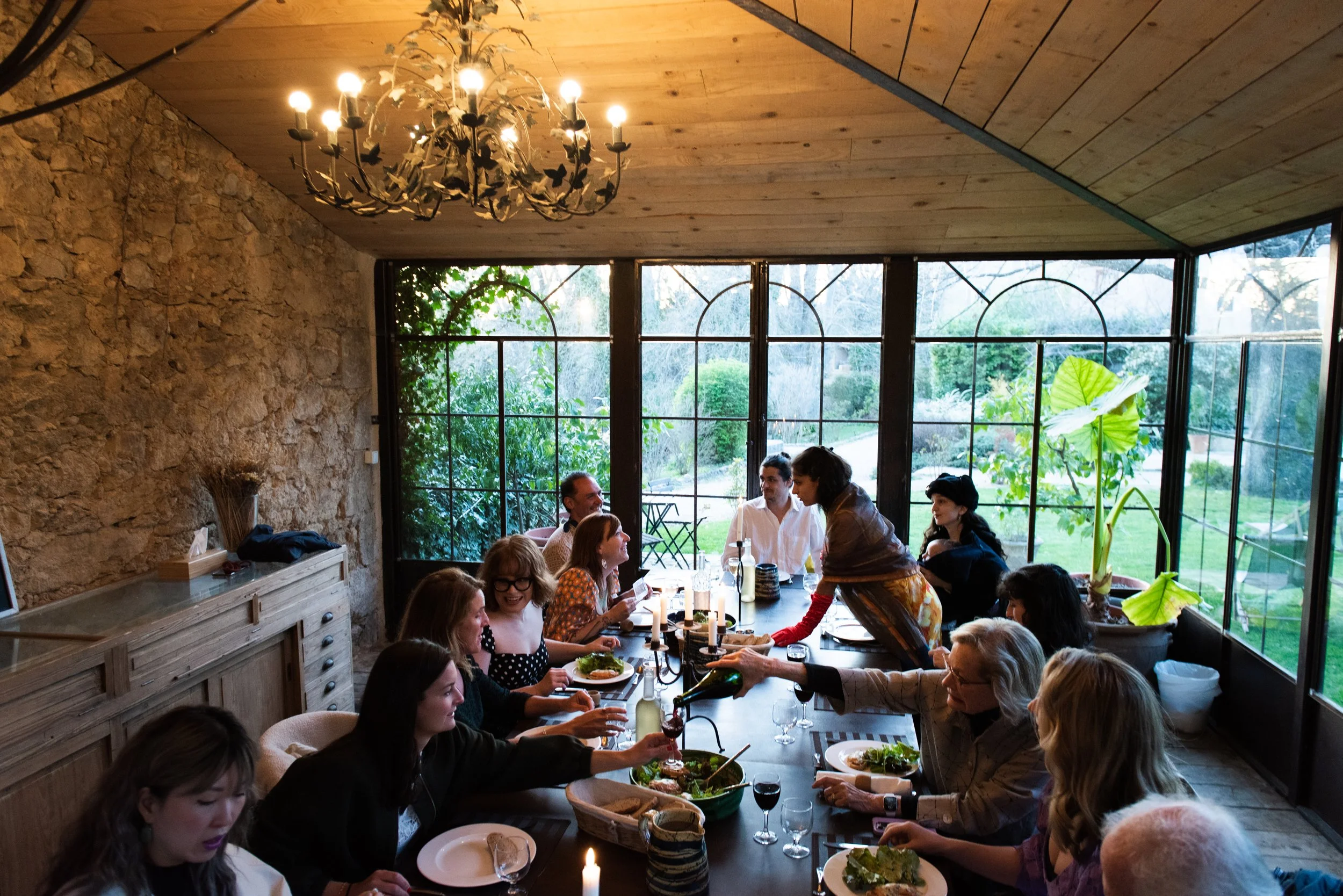 A group of people gathered around a long dining table inside a room with large windows, a stone wall, and a wooden ceiling, enjoying a meal. breathwork events portland