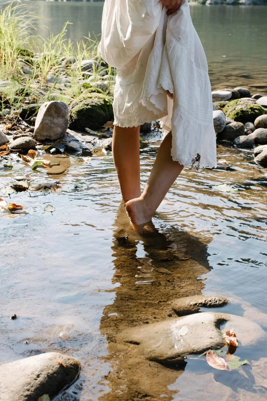 Person standing in shallow water near a rocky shoreline, wearing a white dress with lace details, with feet touching the water and surrounded by stones and greenery. trauma-informed breathwork Portland Oregon