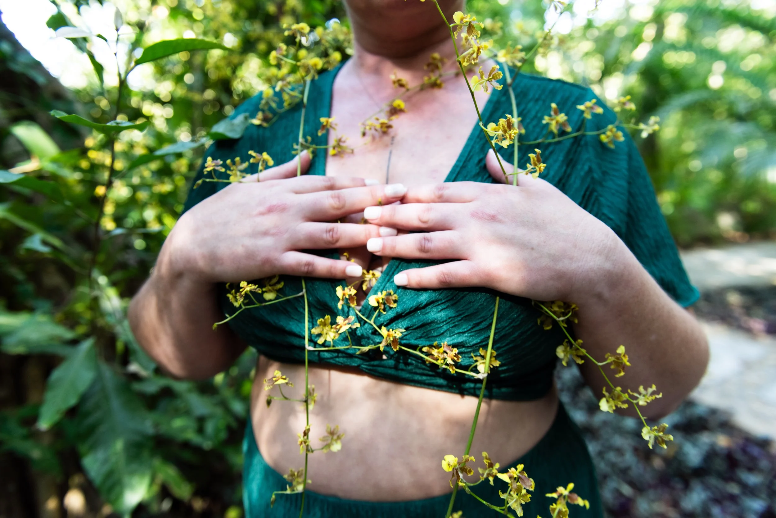 A person with a bare chest wearing a green top amidst green foliage, with yellow flowers covering part of their body and face, touching their chest. retreat photographer