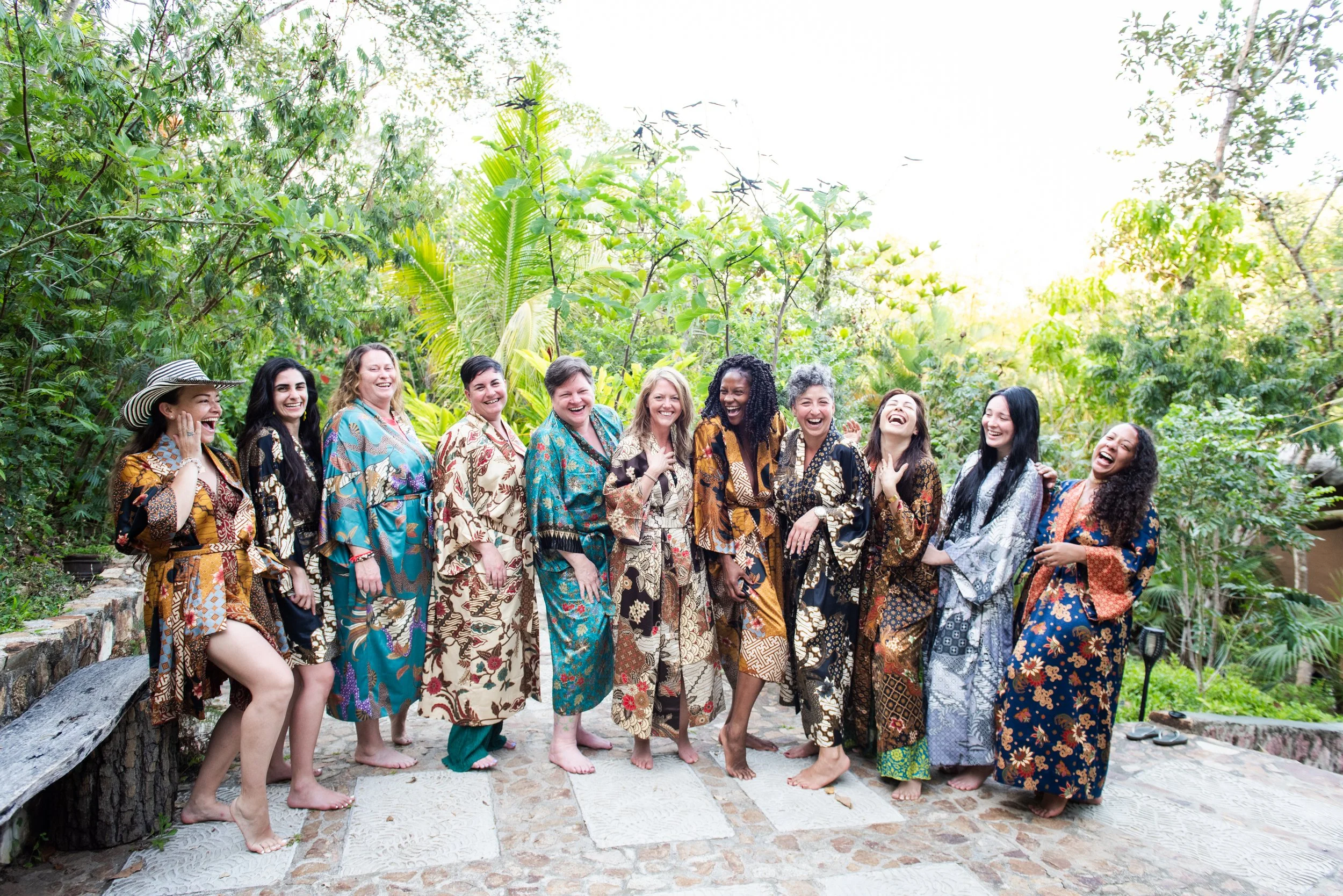 A group of women dressed in colorful, patterned dresses standing and laughing together outdoors in a lush, green garden or jungle setting. international retreat photographer