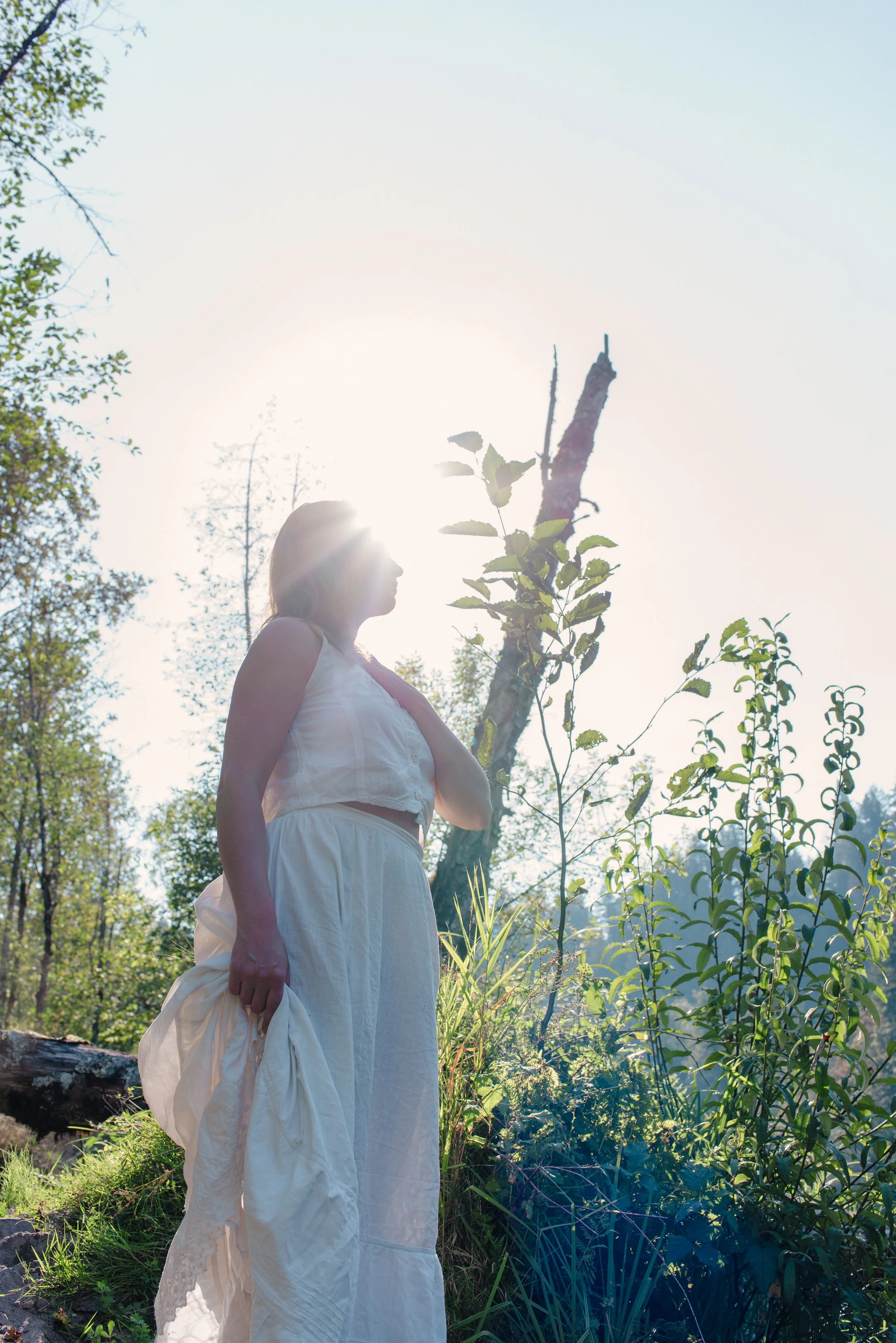 Trauma Informed Breathwork Guide Briana Lynn Cerezo in a white dress standing outdoors in a wooded area with sunlight shining behind her, surrounded by trees and greenery.