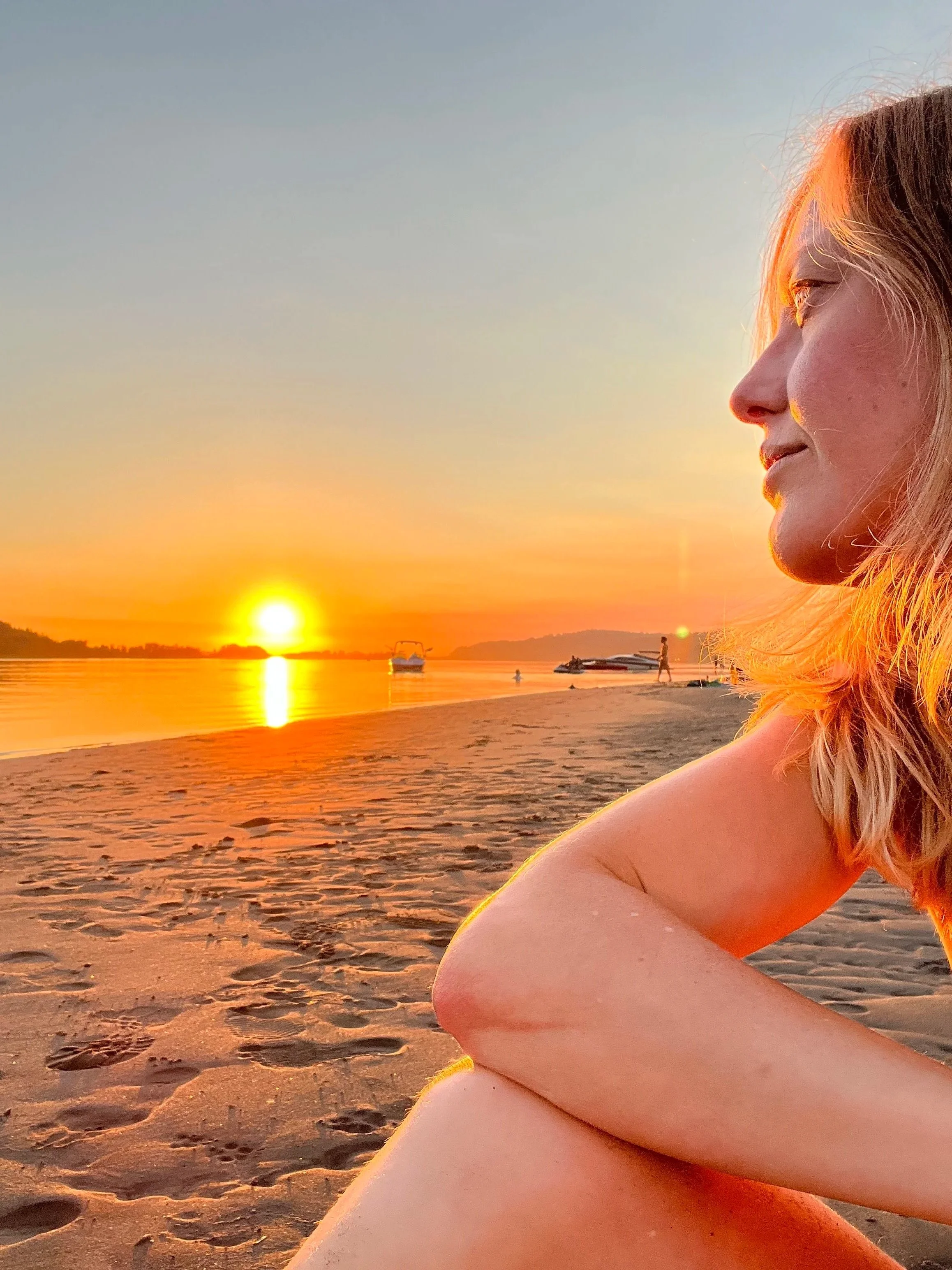 Close-up side profile of Briana Lynn Cerezo sitting on the sandy beach, enjoying the sunset over the water with boats and a person walking in the distance. Gentle breathwork portland