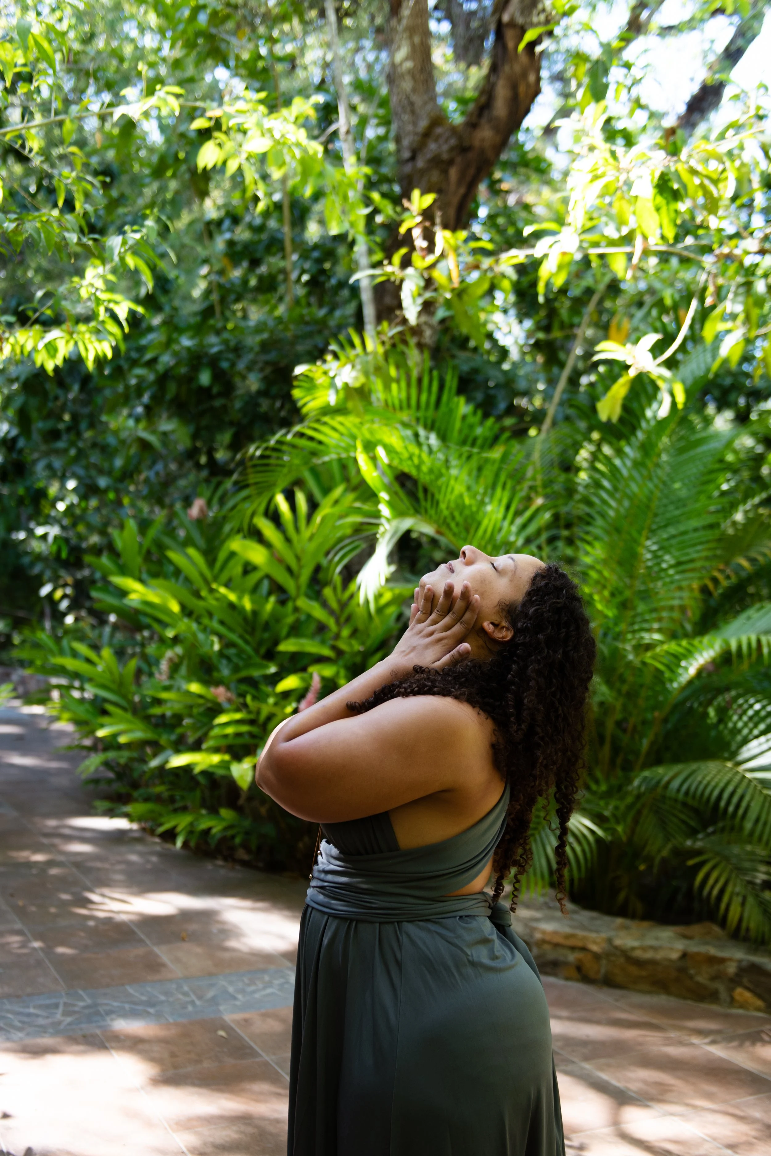 A woman with curly hair standing outdoors among lush green plants, wearing a dark grey wrap dress, and holding her face with both hands in a serene pose. sacred portrait photographer