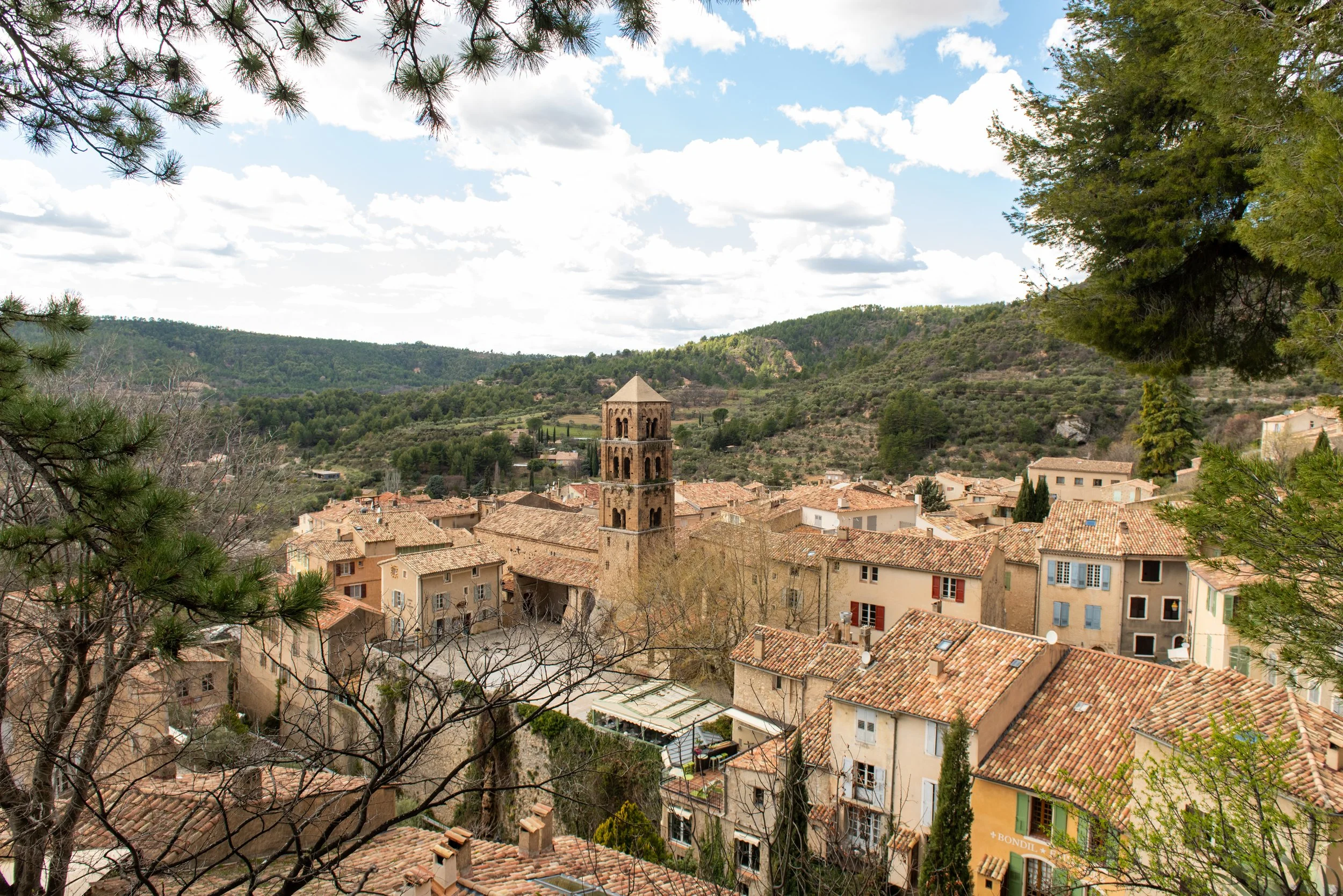 A scenic view of a small town with a brick tower church in the foreground, surrounded by terracotta rooftops, green hills in the background, and partly cloudy sky above, framed by pine tree branches. breathwork portland