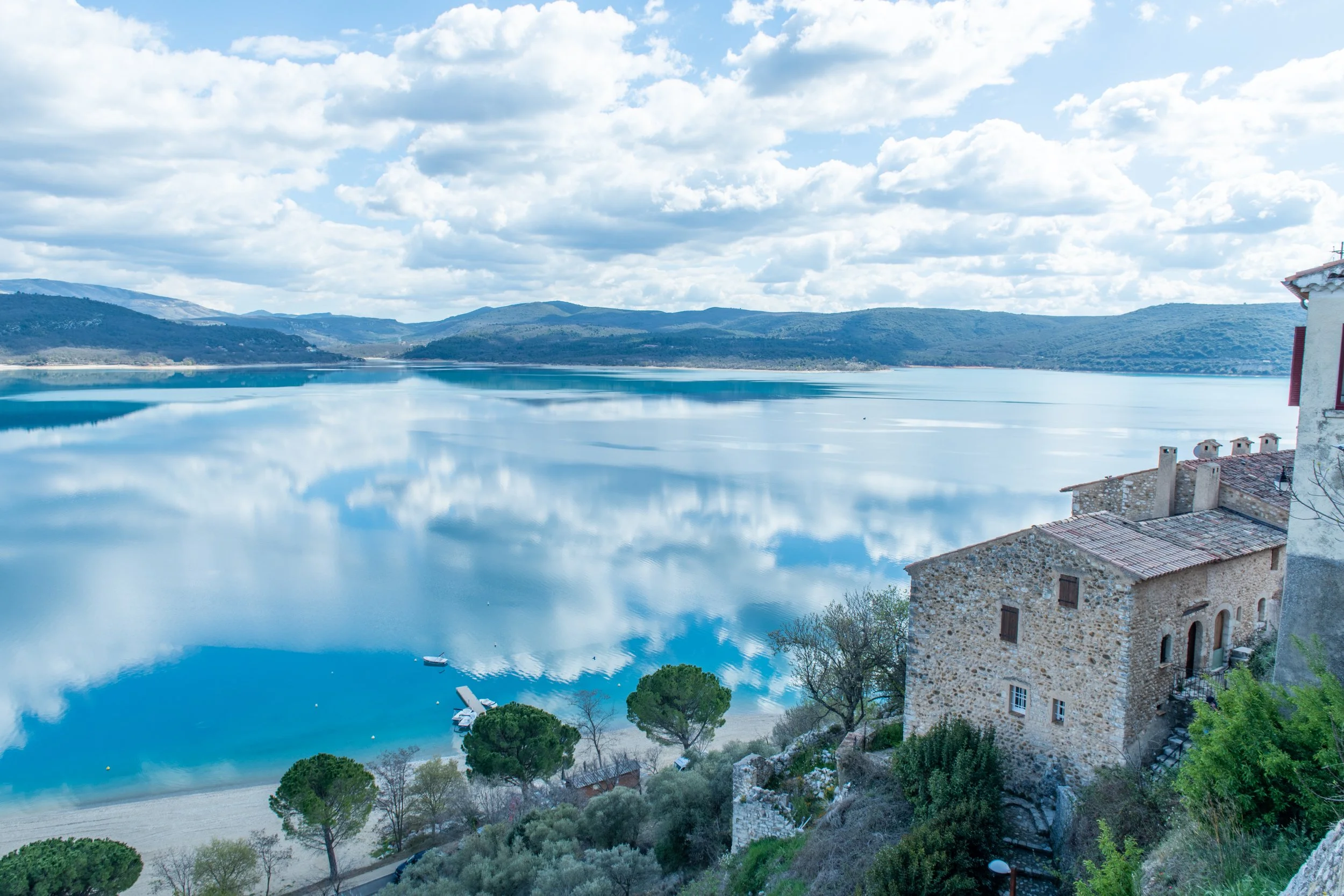 A serene lake surrounded by green hills and mountains under a partly cloudy sky, with reflections in the water and a small boat near the shoreline next to an old stone building. breathwork ceremony portland