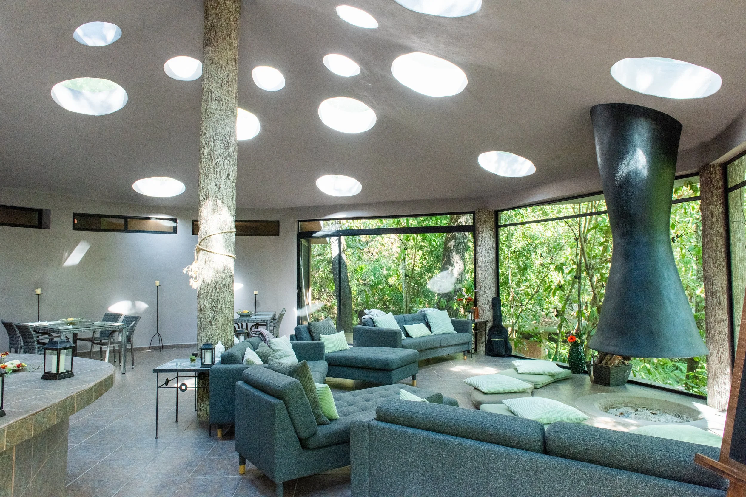 Indoor living space with large windows, blue sofas with green and patterned pillows, a dining area with tables and chairs, a tree trunk in the middle, and a ceiling with circular skylights allowing natural light, surrounded by lush greenery outside. 