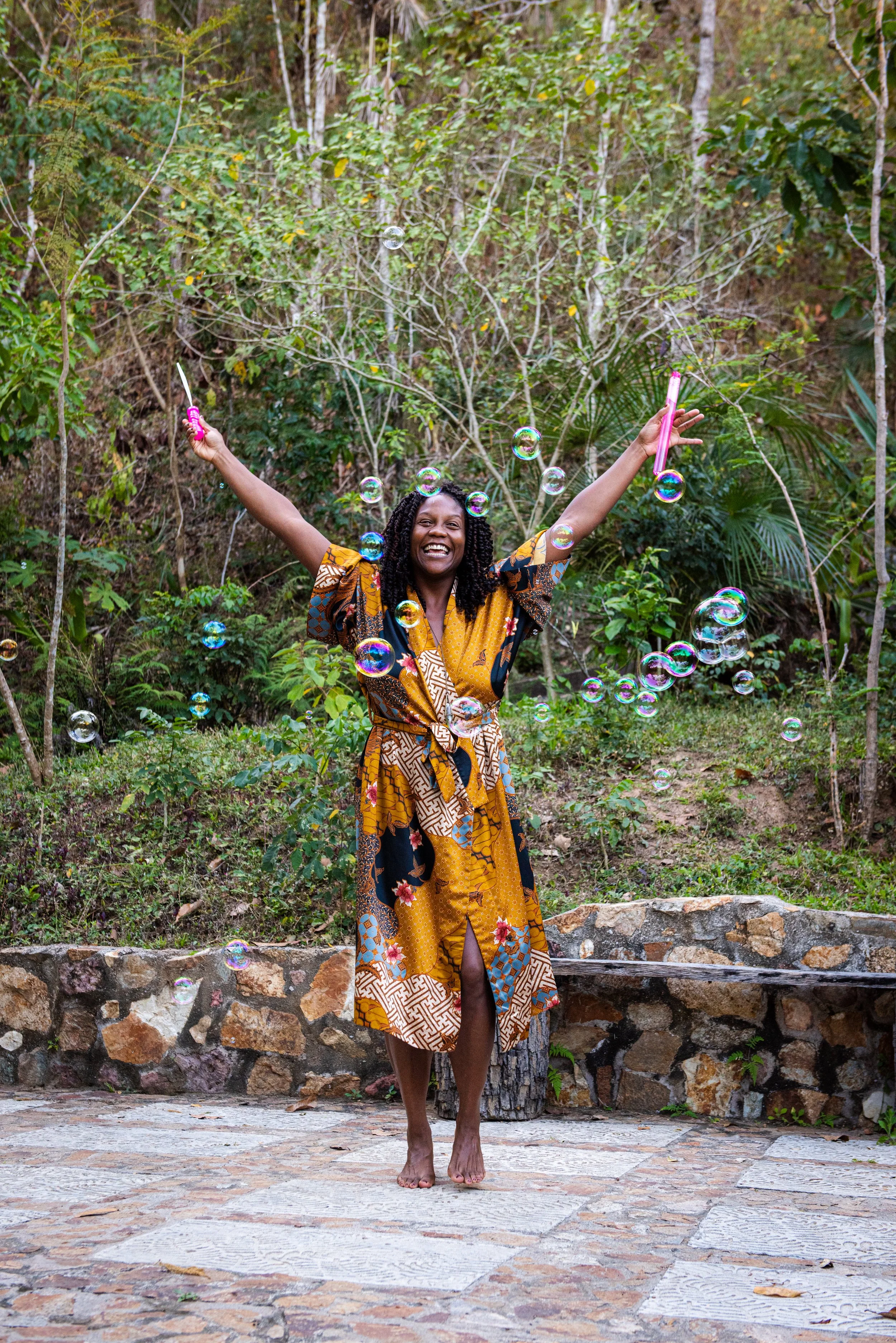 A woman wearing a colorful patterned dress stands barefoot on a stone patio, surrounded by greenery and trees, joyfully tossing soap bubbles into the air. sacred portrait photographer portland
