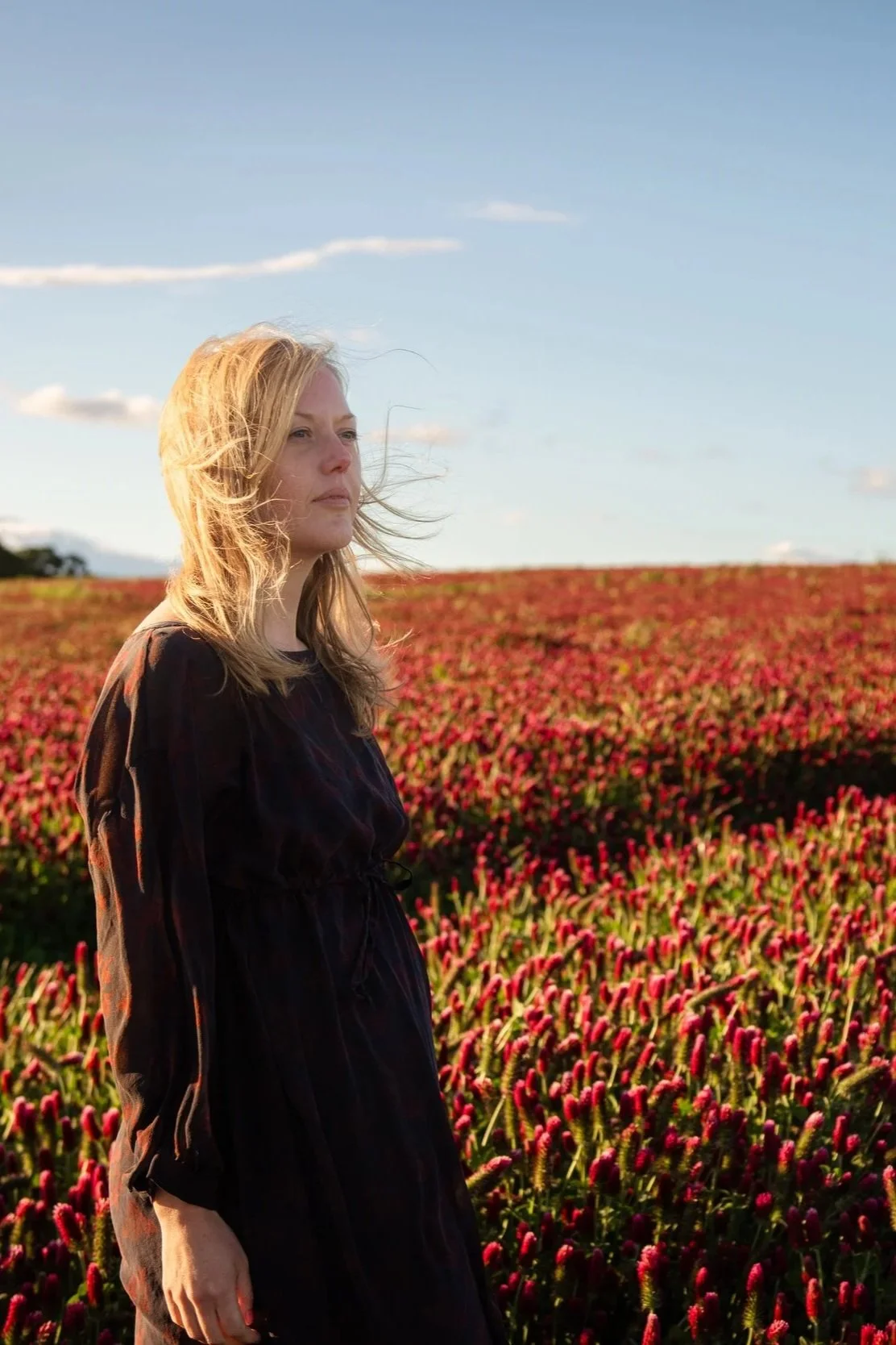 Briana Lynn Cerezo standing in a field of pink flowers under a blue sky with scattered clouds. Breathwork guide portland