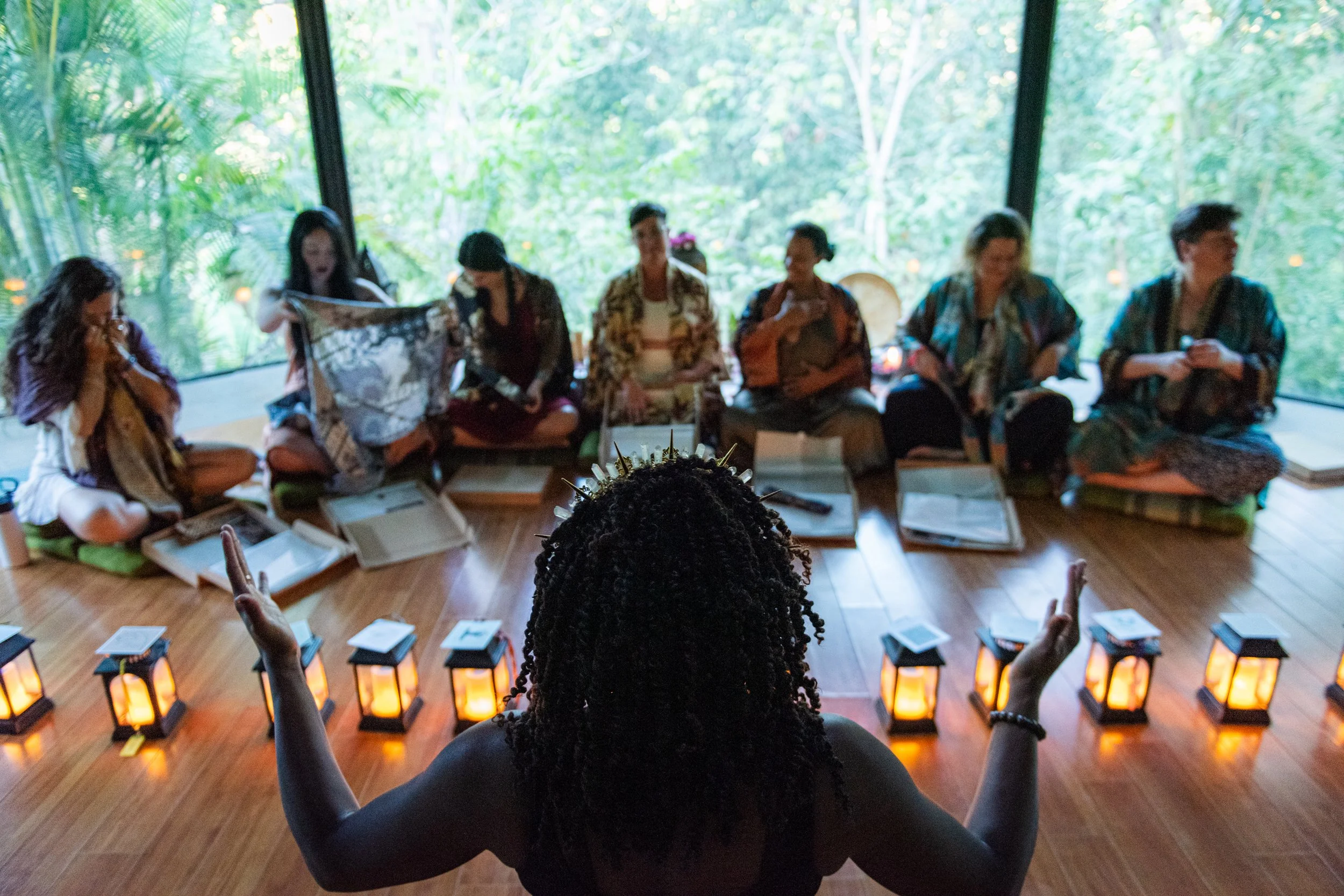A woman with braided hair sitting with arms raised in front of a group of women in a circle, engaged in a gathering with candles and books, with lush green foliage visible through large windows. breathwork ceremony portland