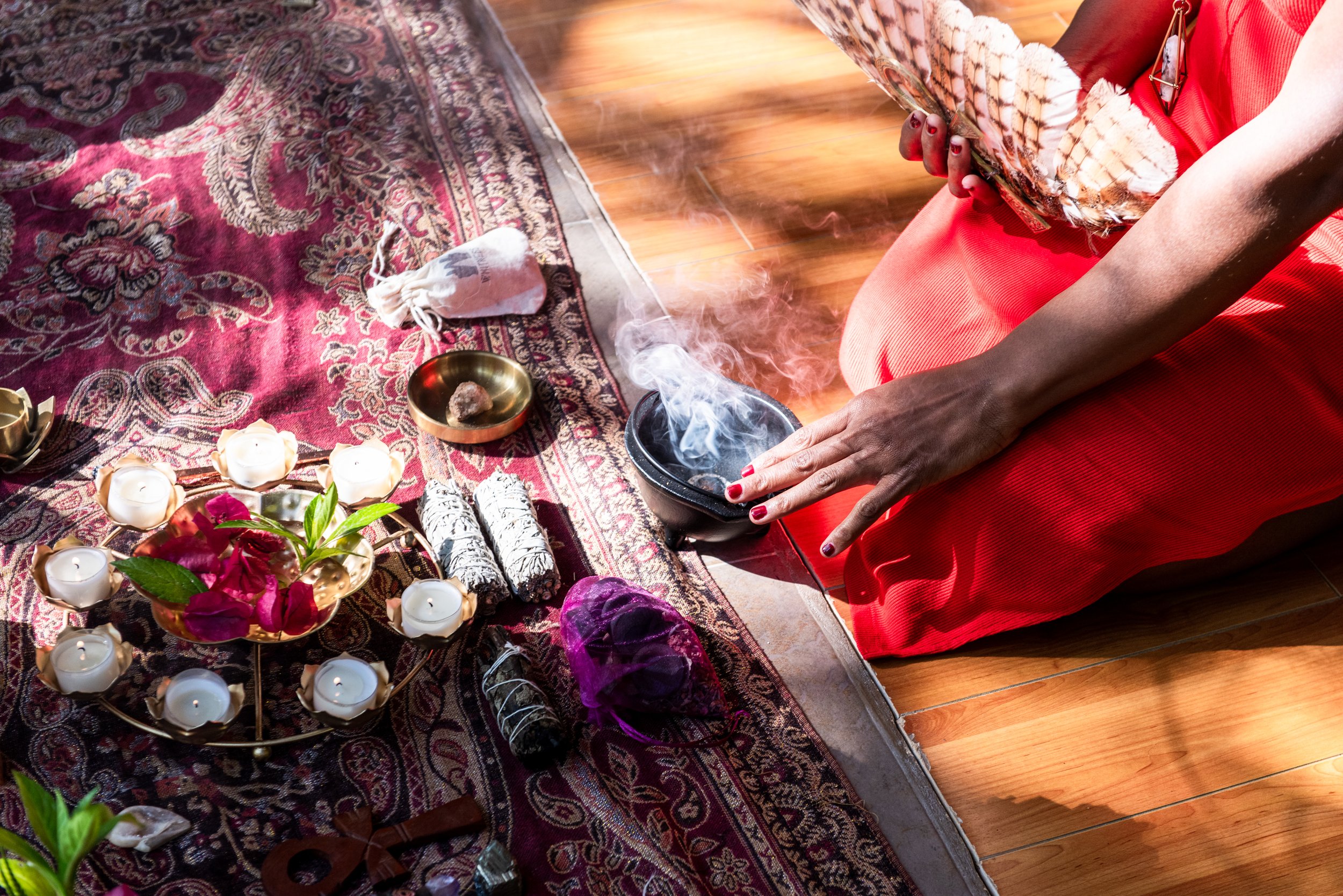 A person in a red dress holding a photo book, sitting on a rug with candles, flowers, and spiritual items in a ritual setting. breathwork guide portland