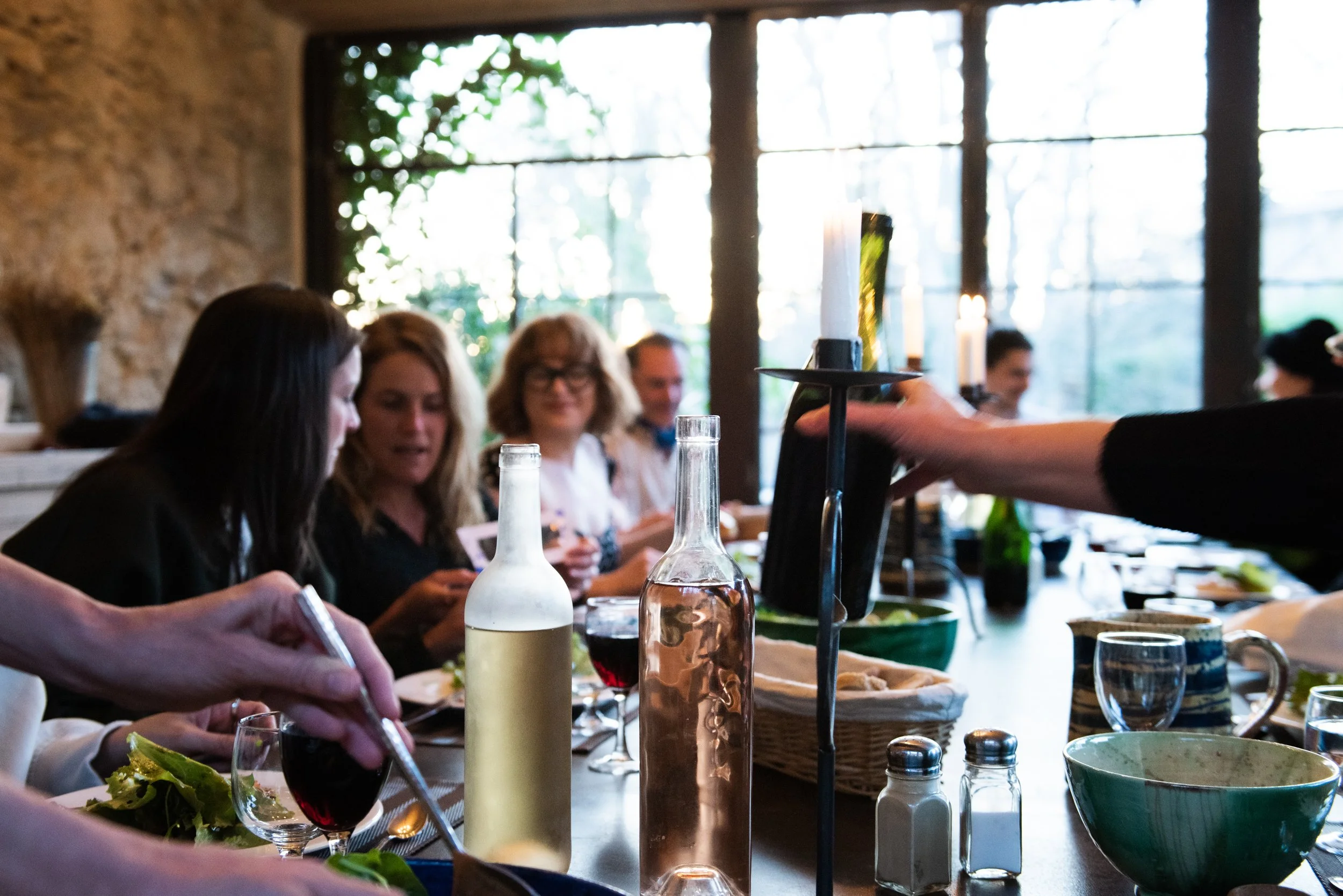 People dining at a restaurant table with bottles and food, with large windows and greenery in the background. breathwork portland