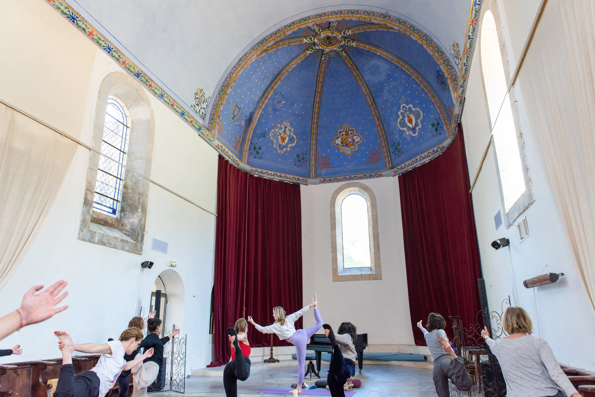 People participating in a yoga or dance class inside a church with blue decorated ceiling, tall arched windows, and red curtains.
