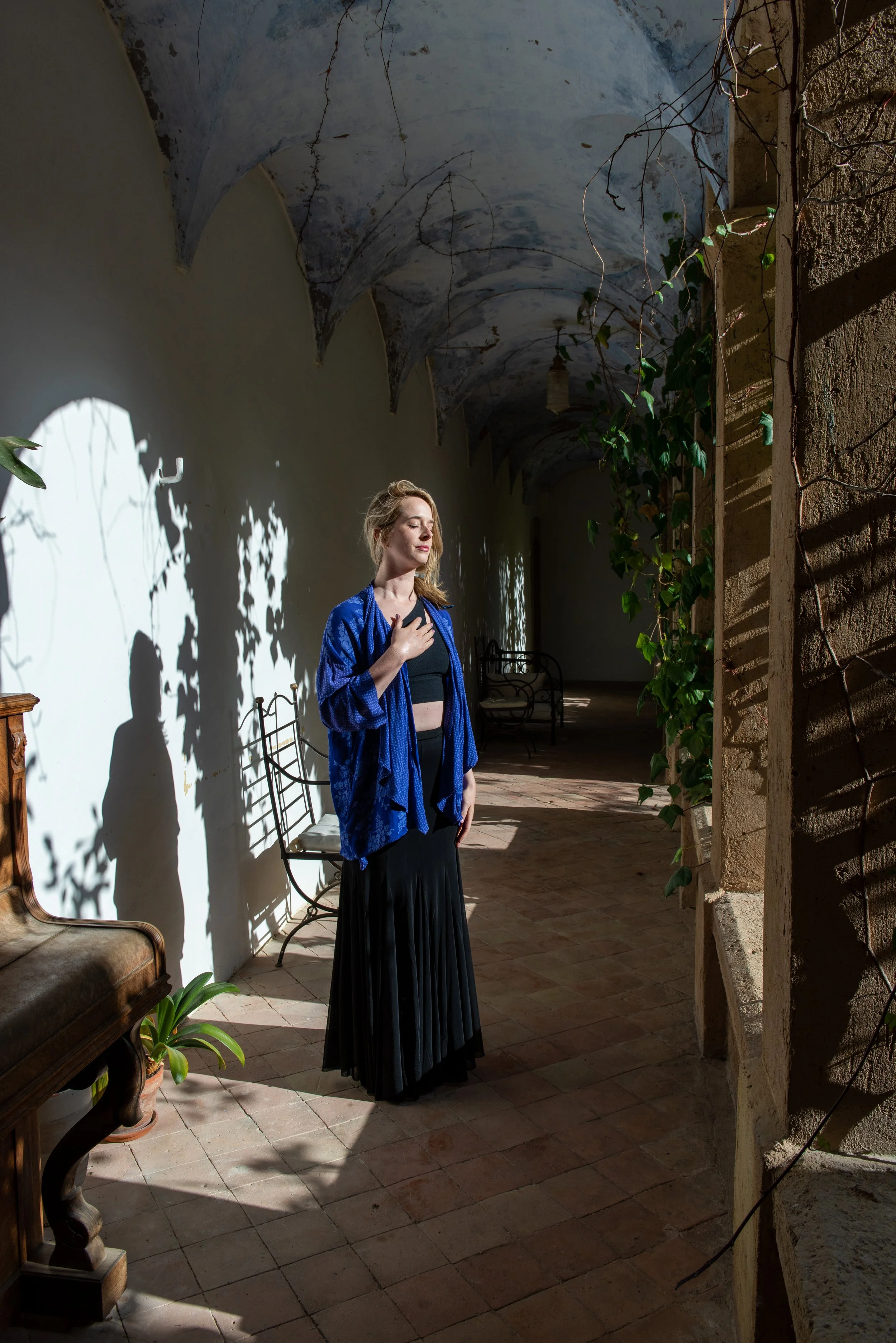 A woman with blonde hair, wearing a blue jacket and a long black skirt, stands in an outdoor corridor with sunlight and shadows, touching her chest with her right hand, with chairs and plants around. nervous system healing portland