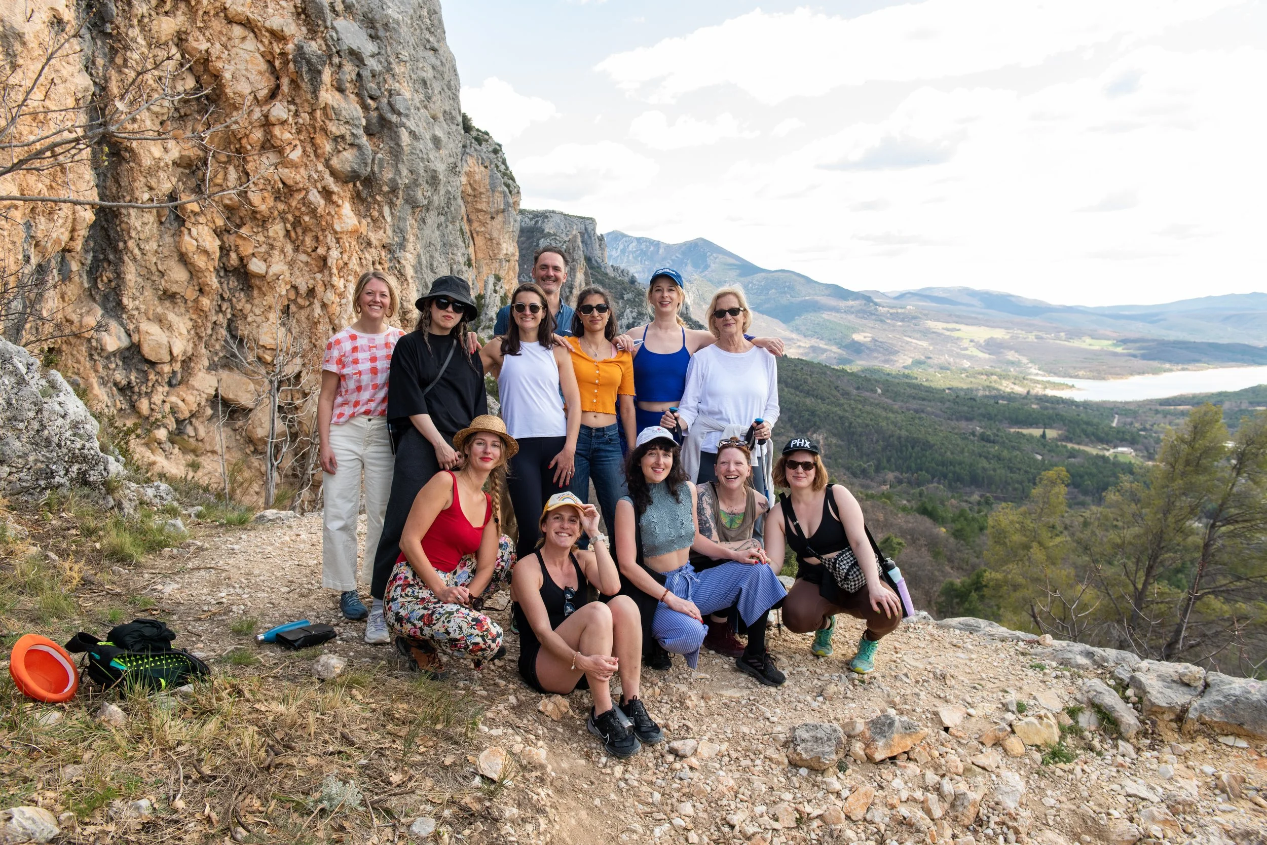 A group of 14 people posing outdoors on rocky terrain with mountainous landscape in the background. They are dressed casually, some wearing hats, sunglasses, and athletic clothing. Briana Lynn Cerezo trauma informed breathwork