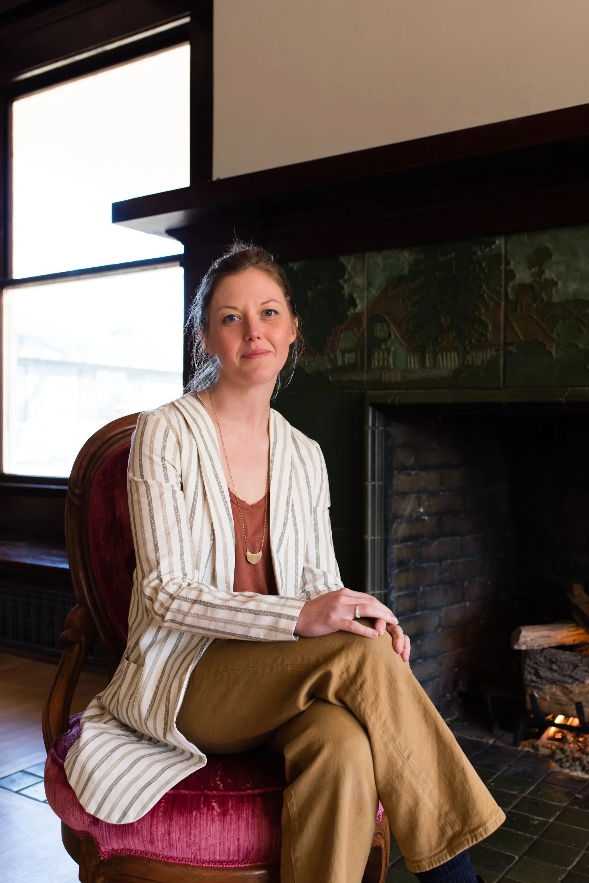 Somatic Integration coach Briana Lynn Cerezo sitting on a vintage red velvet armchair by a fireplace in a cozy room, with a framed painting of a house and trees on the wall behind her.