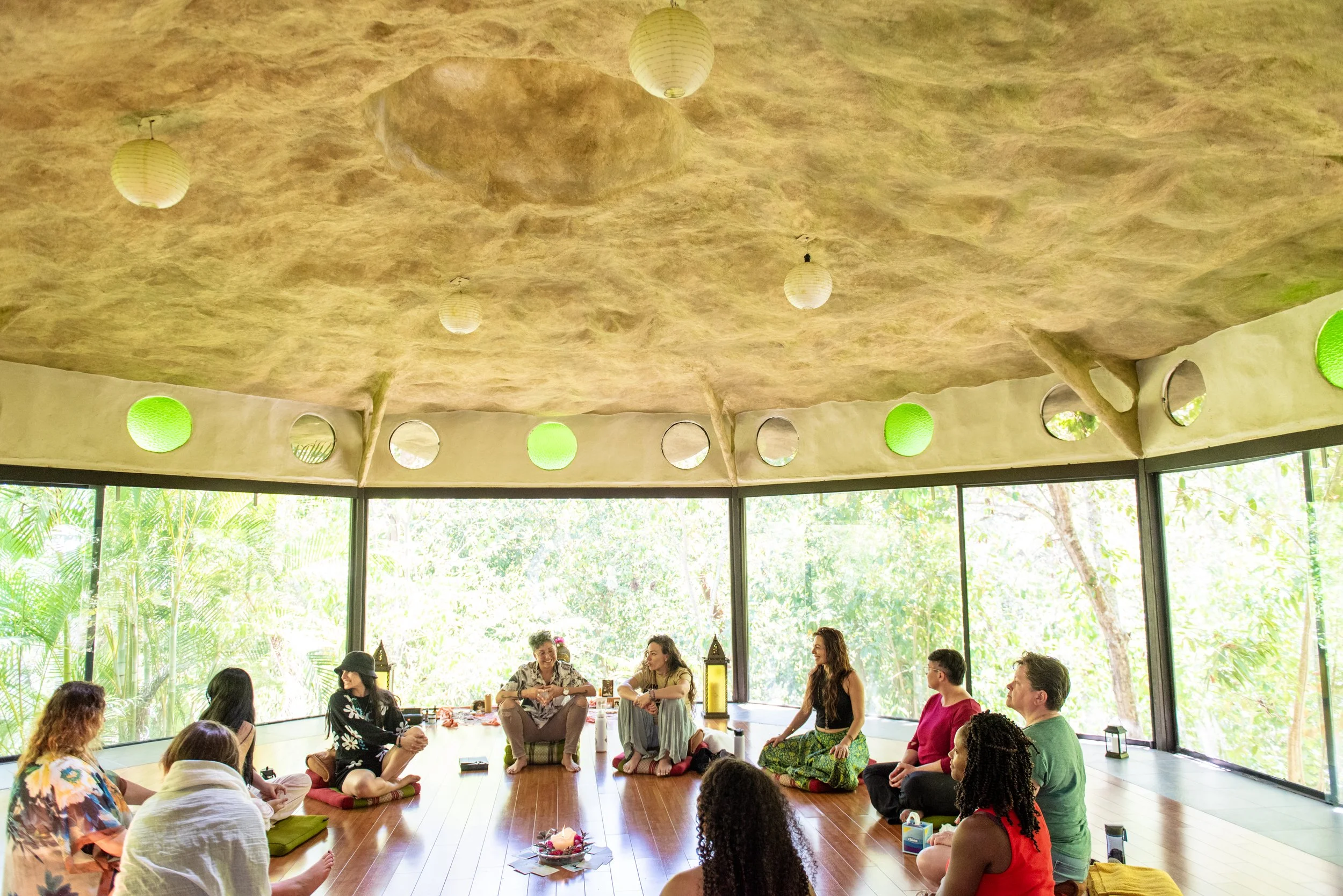 People sitting in a circle inside a room with large windows and a natural environment outside, engaging in a group discussion or meditation, with a decorative arrangement in the center. wellness retreat facilitator