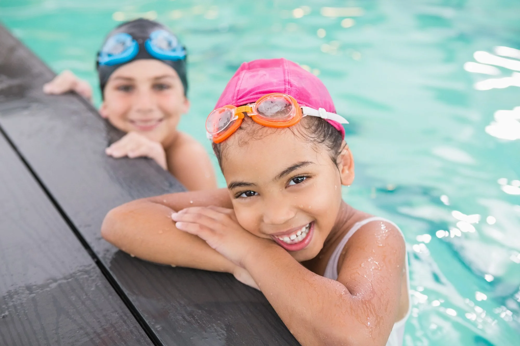  Two little girls wearing swim caps and goggles in a swimming pool with arms leaning over the edge of the pool smiling at camera. 