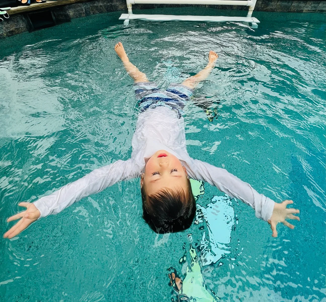  Young boy around 4 years old floating on his back on the the top of the water with his legs and arms out stretched. He is wearing a white long sleeve shirt with blue stripped shorts. 